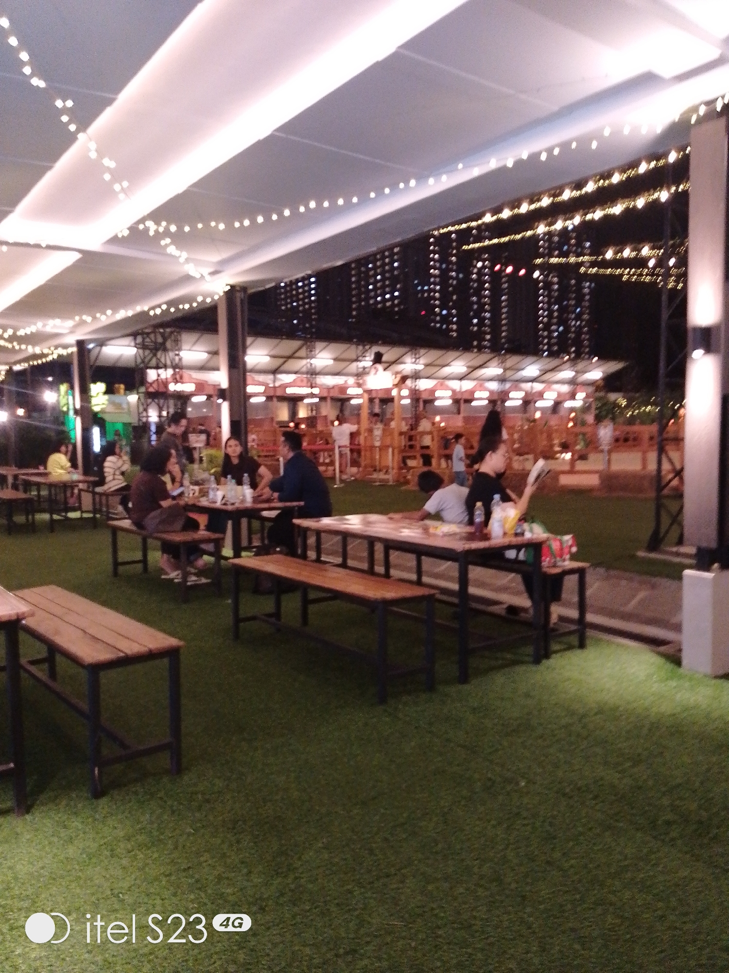 The image depicts an outdoor seating area at night, likely a restaurant or cafe, in Surabaya, Indonesia. String lights are draped across a white canopy, illuminating the scene. Several groups of people are seated at wooden tables with benches. In the foreground, empty wooden tables and benches are set on an artificial grass lawn. In the mid-ground, a group of individuals are gathered around a table, with some engaged in conversation and others looking at their phones. Further back, more tables and patrons are visible. In the background, the lights of a city skyline, likely Surabaya, can be seen. The overall atmosphere is casual and social, with the lighting suggesting a festive evening setting.