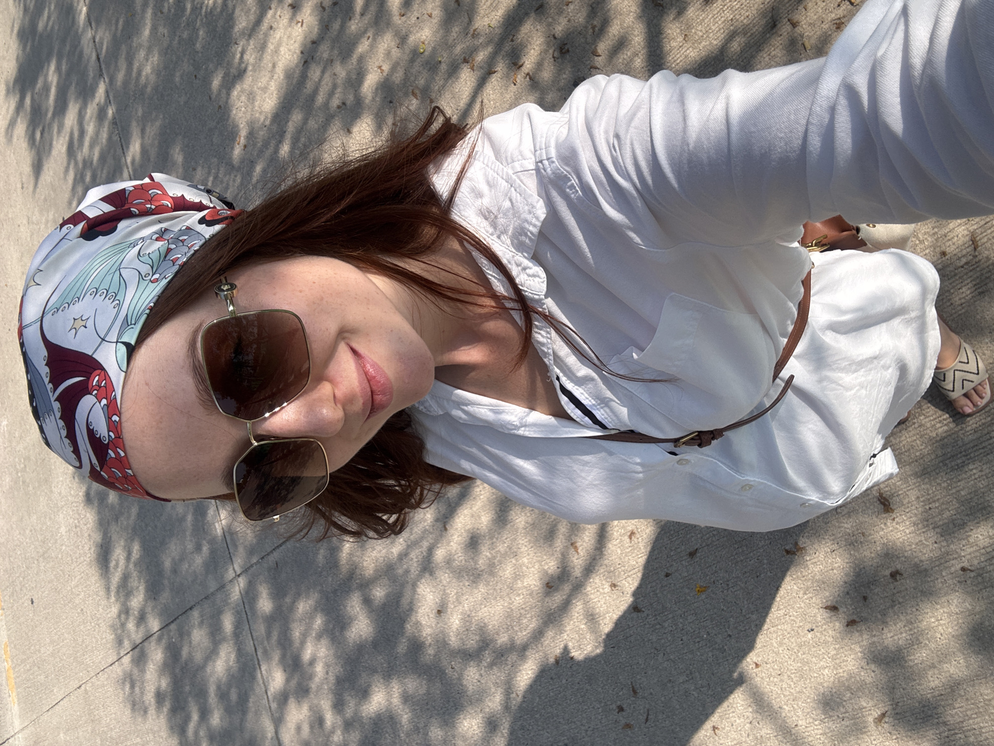 An overhead, slightly tilted view captures a person, likely a woman, outdoors on a sunny day. She looks upwards with a subtle smile. Her head is covered with a stylish headscarf in light blue, red, white, and dark blue/purple tones, featuring an intricate design that resembles mythical creatures or elaborate patterns. She wears fashionable sunglasses with dark lenses and gold-toned frames.
Her attire includes a long-sleeved, collared white shirt dress made of a light fabric, belted at the waist with a slim, dark brown leather belt featuring a small gold buckle. A glimpse of a dark striped undergarment is visible beneath the dress. On her feet, she wears light-colored sandals with a patterned strap, possibly in a zigzag design.
The setting is an outdoor paved area, likely concrete, brightly lit by the sun. The ground is covered with distinct, dappled shadows from overhead tree foliage, and scattered dry leaves are visible. The strong sunlight and sharp shadows suggest it is midday or early afternoon. The overall scene conveys a relaxed atmosphere in a warm climate, consistent with the location context of Choeng Thale, Thailand. No visible text is present in the image.