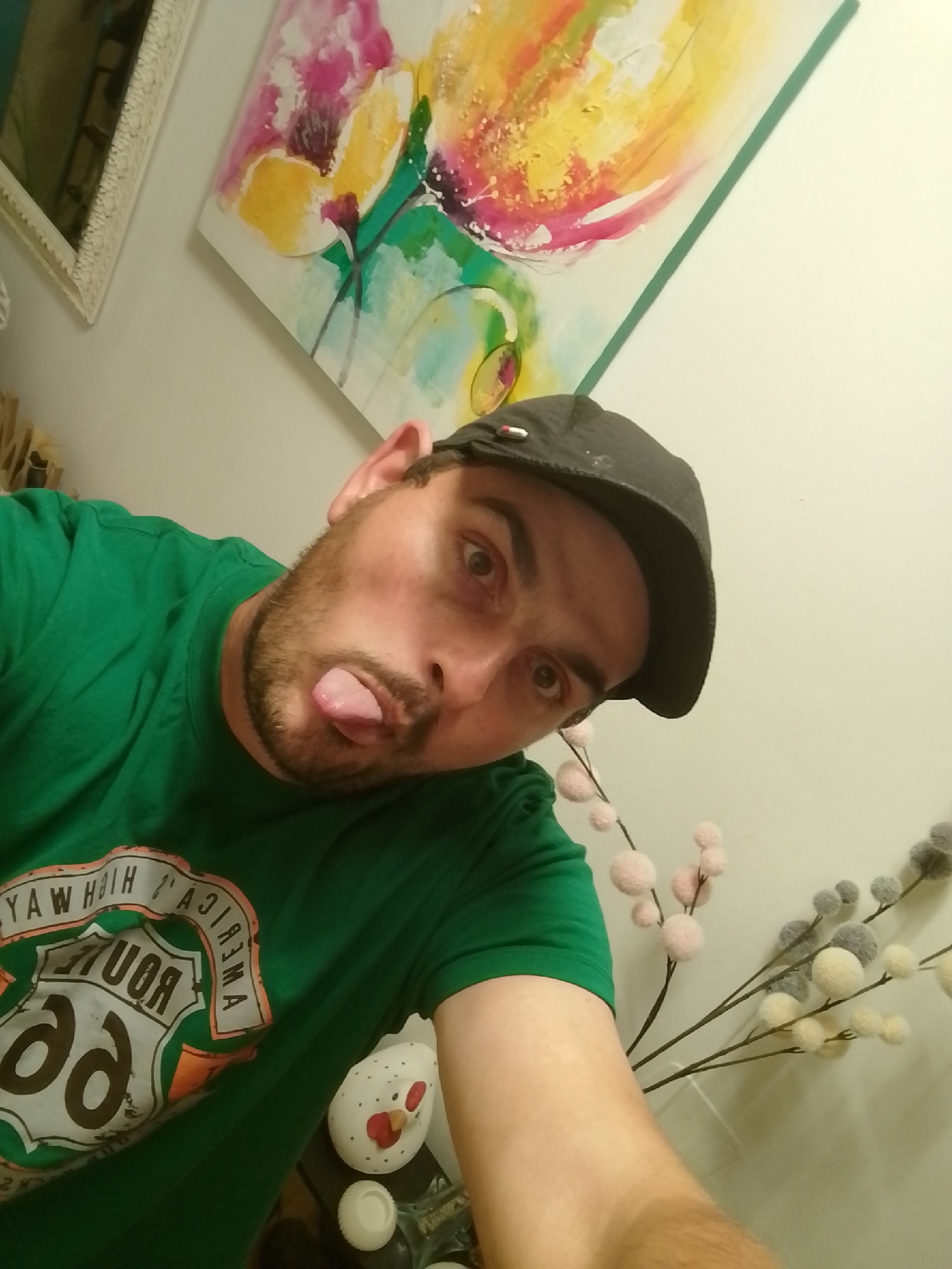 The image is a selfie of a man in Wedel, Germany, taken indoors. The man is wearing a green t-shirt with a Route 66 logo and a dark baseball cap. He is sticking his tongue out at the camera. In the background, there is a large, colorful abstract painting on the wall and a decorative branch with fuzzy pink and grey balls. A white ceramic chicken figurine is also visible on a surface to the right. The lighting suggests it is daytime, and the setting appears to be a home or a casual indoor space.