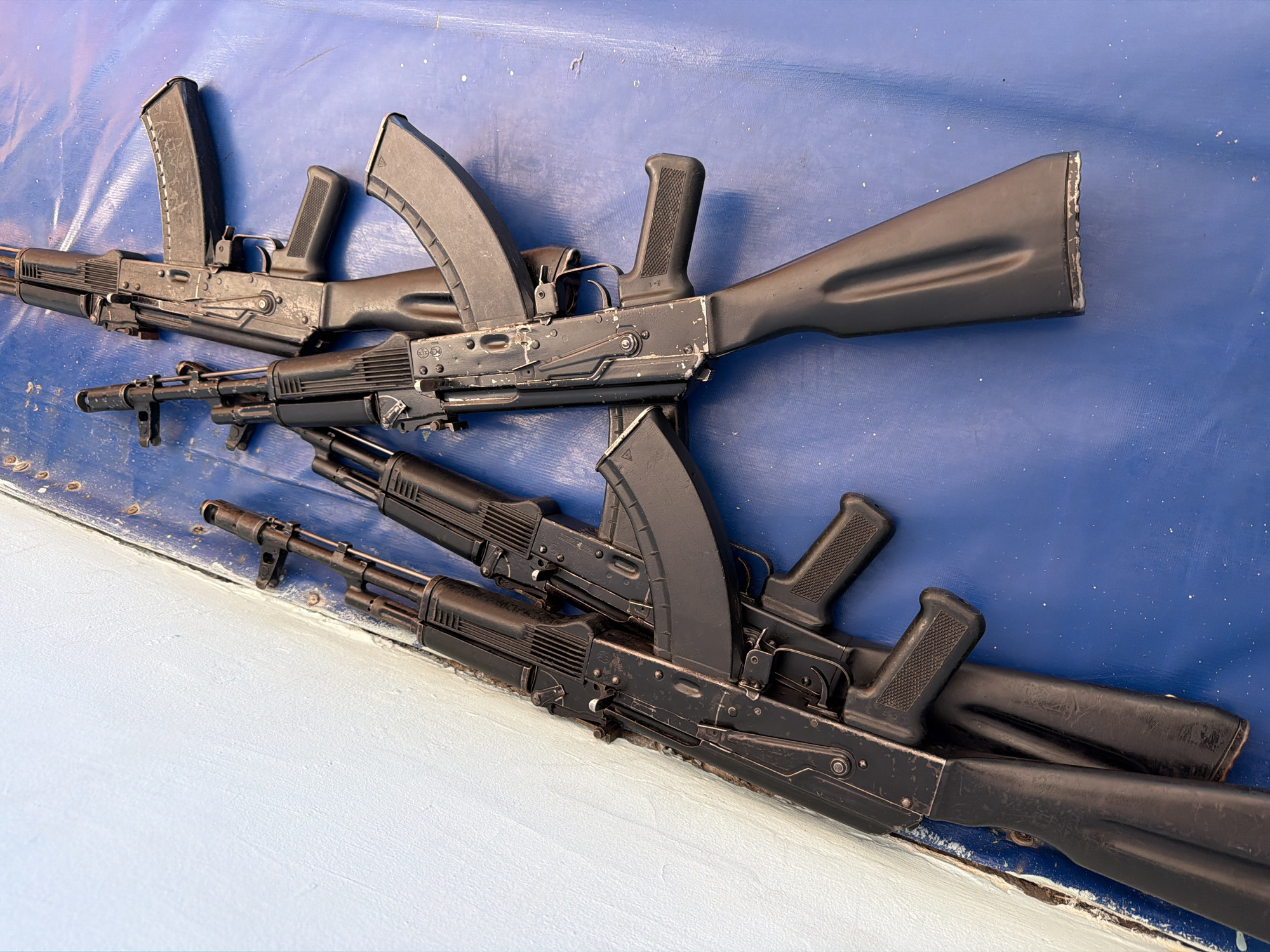 The image shows four black AK-pattern rifles with curved magazines lying on a light blue and white painted surface. The rifles appear to be used, with visible wear and tear. The rifles are arranged in a slightly overlapping manner. There are no people visible in the image. The specific location in Izhevsk, Russia, is not identifiable from the image.