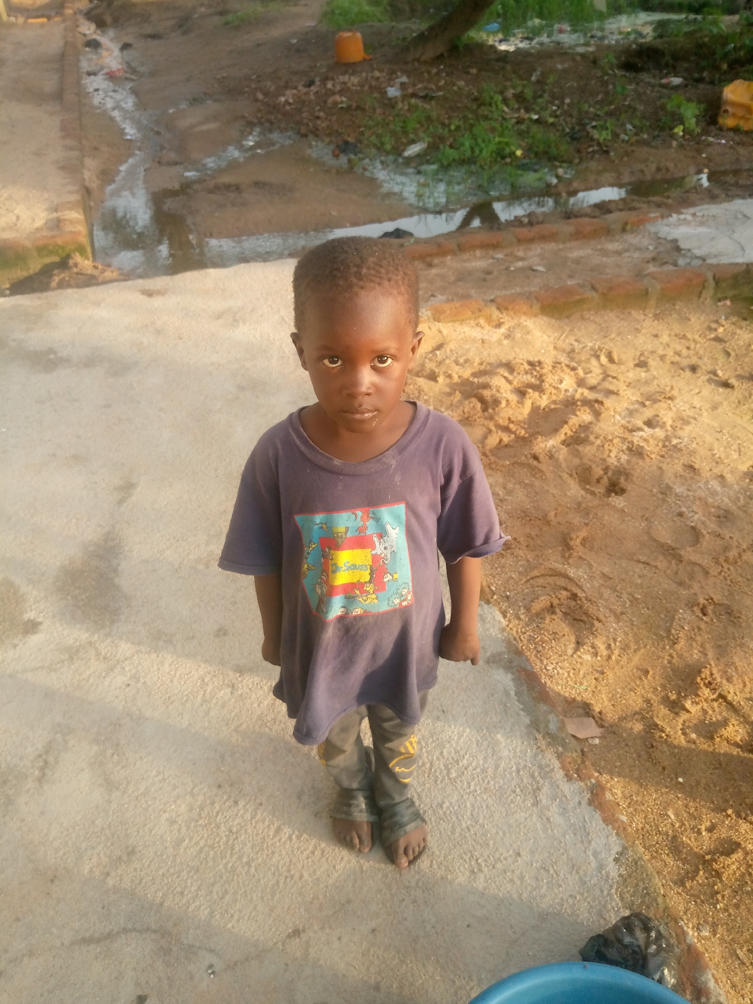 A young boy stands on a concrete pathway in Jauro Yinu, Nigeria, looking directly at the camera. He is wearing a dark blue t-shirt with a colorful graphic on the front and dark pants. The boy's feet are bare. To his left, the concrete pathway is wet and has a small stream running along it, bordered by bricks. Beyond the bricks, there is a patch of sandy soil with scattered debris and vegetation. To his right, the sandy soil extends further, with evidence of footprints. In the background, there is more vegetation and a yellow plastic container. The lighting suggests it is late afternoon, with shadows cast by the sun.