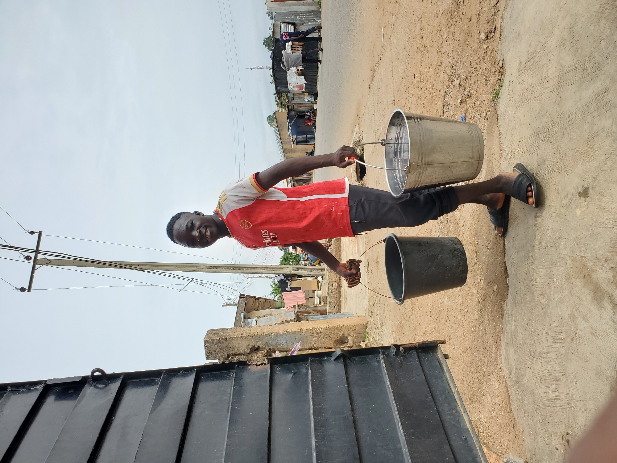 A man in a red and white shirt and black pants carries a black bucket and a silver bucket, one in each hand. He stands on a concrete surface beside a dirt road, smiling at the camera. In the background, there are buildings and a street. The scene is likely located in Jos, Nigeria. A black metal gate is at the bottom left of the image. The sky is overcast.