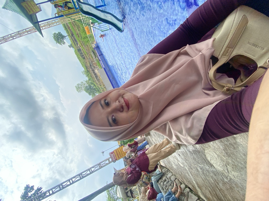 A woman in a pink hijab and maroon top poses beside a pool with a water slide. She's holding a beige purse with "Elizabeth" written on it. Behind her is a cloudy sky. Some other people are sitting on a stone surface visible at the bottom. A structure with metal beams stands to the left and a water park or recreational area is in the background.