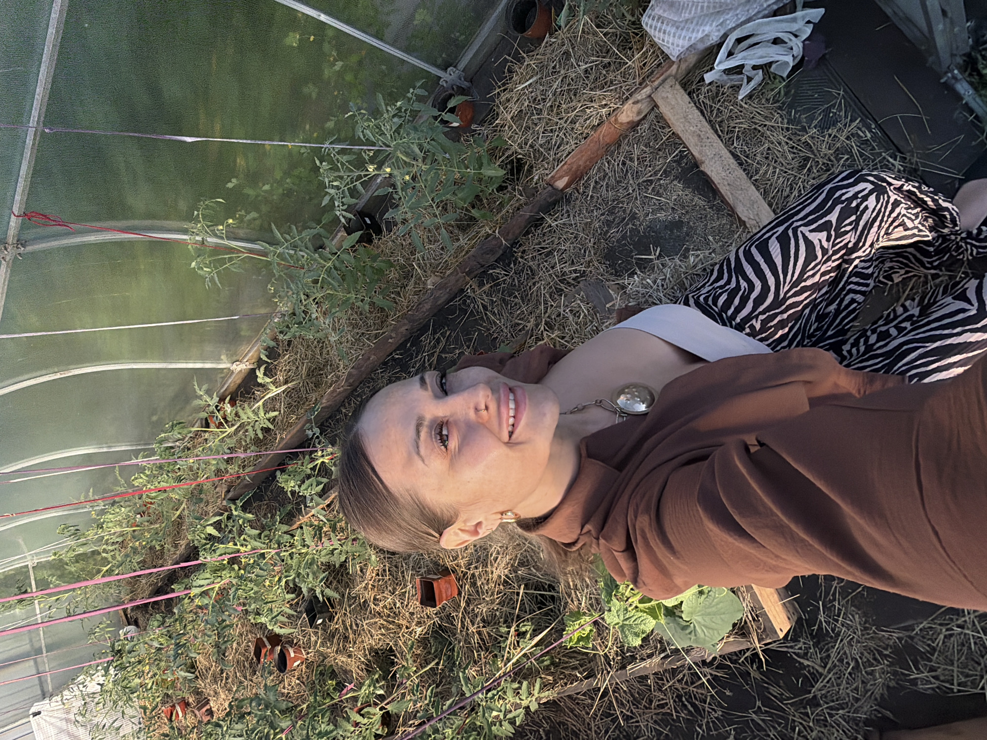 In a greenhouse in Kansk, Russia, a woman with brown hair pulled back in a ponytail poses for a selfie. She is wearing a brown open-collar shirt, a white top, and black and white zebra-print pants. Her left arm is extended, holding the camera. Her right hand is visible near her lap. A large, round, silver pendant hangs from a chain around her neck. The greenhouse is filled with rows of tomato plants, supported by stakes and strings. The ground is covered in straw. Wooden planks form the raised beds for the plants. The greenhouse structure is visible in the background, with translucent panels allowing light to enter.