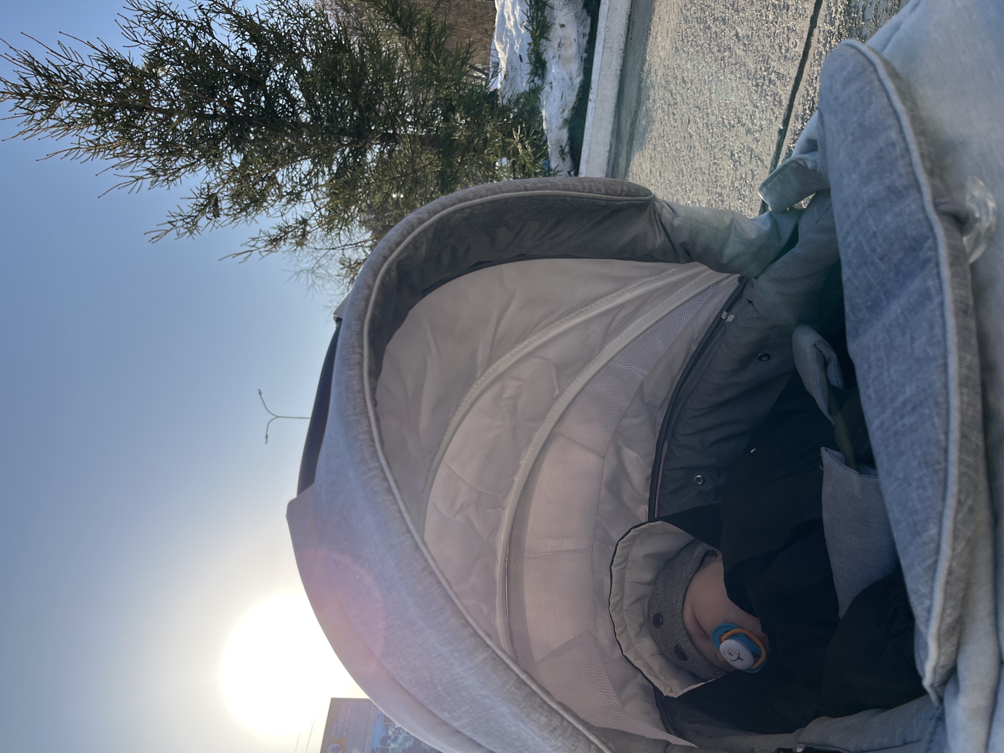 The image displays an infant asleep in a light grey fabric stroller. The infant is wearing a dark garment and a grey knit hat with two dark circular details, with a blue and orange pacifier in its mouth. The stroller's canopy is partially extended, revealing internal mesh sections. The scene is outdoors under a clear blue sky with the sun low on the horizon in the upper-left, causing lens flare. In the mid-ground to the right, a tall, dark green coniferous tree is prominent, with patches of snow visible on the ground beneath it and extending into the background. The ground directly adjacent to the stroller appears to be paved. Two streetlights on a pole are visible against the sky in the upper-middle background. A large billboard with partially discernible Cyrillic text, including "ТВОЕМУ", is located in the far-left background. The environment is consistent with an urban or park setting in Chaikovsky, Russia, during cold, sunny weather.
