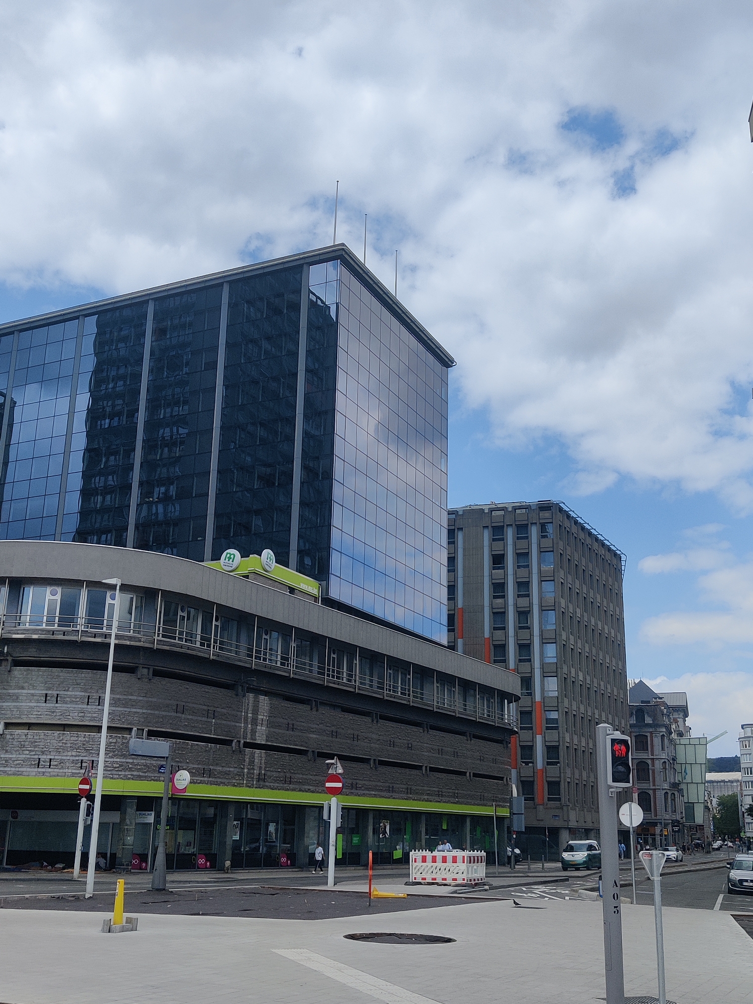 The image depicts a street scene in Liège, Belgium, characterized by modern and older architecture. In the foreground, a pedestrian area is visible with a light gray pavement and some construction barriers. A black bird is standing on the pavement. Further back, there are several buildings. The most prominent is a large, contemporary building with a reflective glass facade, towering over other structures. To its right, a more utilitarian, gray concrete building with numerous windows stands. In the mid-ground, a curved structure with a gray facade and large windows is visible, with a bright green stripe along its upper edge. Several businesses with green signage are located beneath this curved section.
There are a few people in the scene. Two individuals are seated near the bottom left of the image, near the entrance of one of the businesses. Another person is walking on the sidewalk in front of the businesses. A small dog is also visible walking on the pavement. In the background, traffic can be seen on the street, including cars and a green electric vehicle. A traffic light with a red signal is visible on a pole in the mid-right of the image. The sky is partly cloudy with scattered white clouds against a blue sky. The overall impression is an urban environment with a mix of modern and older architectural styles.