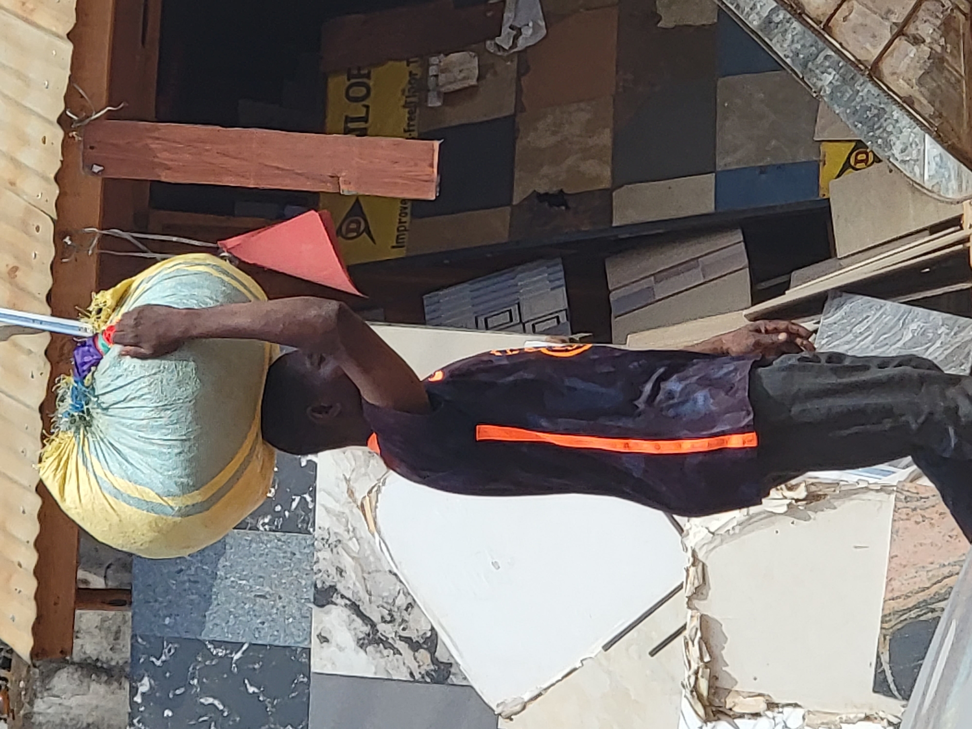 A man in a dark shirt with orange stripes carries a large, yellow and blue sack on his head in Maiburiji, Nigeria. The sack is tied at the top with blue and red string. The man is walking outdoors, with a tiled wall visible behind him. There is a wooden structure overhead, and a red triangular flag hangs from wires. A yellow sign with black lettering, partially obscured, is visible in the background. The man is wearing dark pants. The ground is not visible.