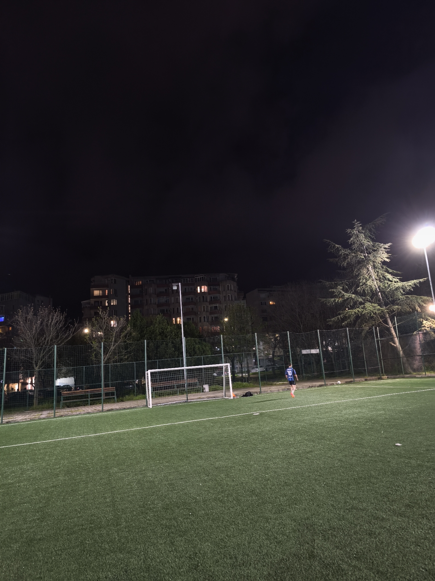 This night-time photograph captures a football (soccer) field with artificial green turf in an urban or suburban setting, specifically in Burgas, Bulgaria. The sky is dark, indicating late evening or night, with the scene illuminated by bright artificial floodlights, one of which is prominently visible on the right side of the frame.
In the midground, a single person, likely a football player, is visible from behind. They are wearing a blue and black jersey with the number "10" clearly displayed on the back, along with orange shorts and light-colored sports shoes. The player is walking across the field towards the right, away from the camera. A white soccer ball rests on the grass near their feet. On the left side of the field, a white football goal with netting stands empty.
The background features several multi-story residential buildings, with numerous windows illuminated, suggesting occupancy. Bare trees and a large evergreen tree are scattered behind the fence that encloses the playing area. Benches are visible behind the goal, indicating a spectator or resting area. The overall impression is of an active community sports facility used after dark.