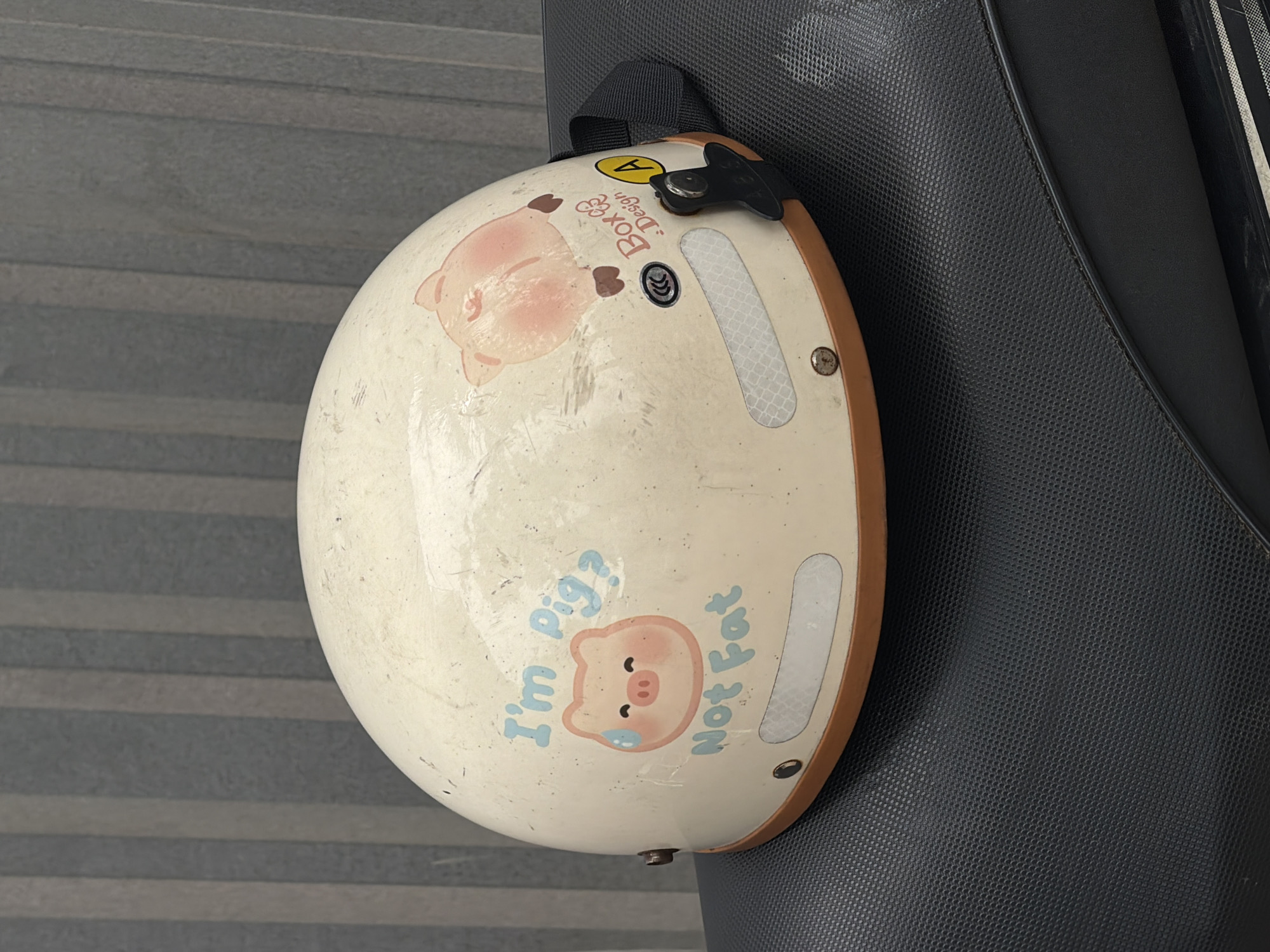 A motorcycle helmet is hanging on the back of a motorcycle seat. The helmet is mostly white with a brown trim and two cartoon pigs printed on it. The top pig is lying on its side while the bottom pig has the words "I'm Pig?" and "Not Fat" printed around it. The background includes a gray concrete wall and the black motorcycle seat. The image was taken in a city, possibly Đà Nẵng, Vietnam.