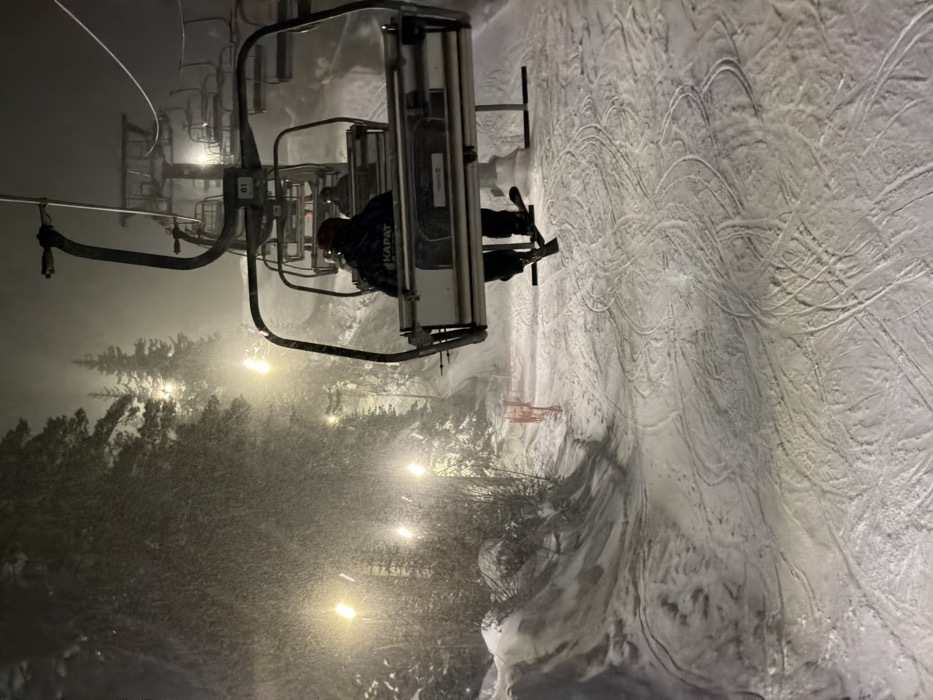 Nechkino, Russia. A nighttime scene depicts a snow-covered ski slope during active snowfall. The environment is illuminated by multiple bright, warm-toned floodlights positioned along the incline, highlighting falling snowflakes and snow-laden coniferous trees that line the left edge of the ascent. In the foreground, an individual is observed from behind, seated in a ski lift chair labeled "01." This person wears a dark jacket with "KAPAT" (Cyrillic script) visible on the upper back, black trousers, and a dark red helmet. Their skis are attached to their boots and hang directly beneath the chair. Two additional individuals are partially visible in the ski lift chair immediately ahead. The snow-covered ground below the lift exhibits numerous distinct ski tracks. A section of orange safety netting is visible on the left side of the slope.