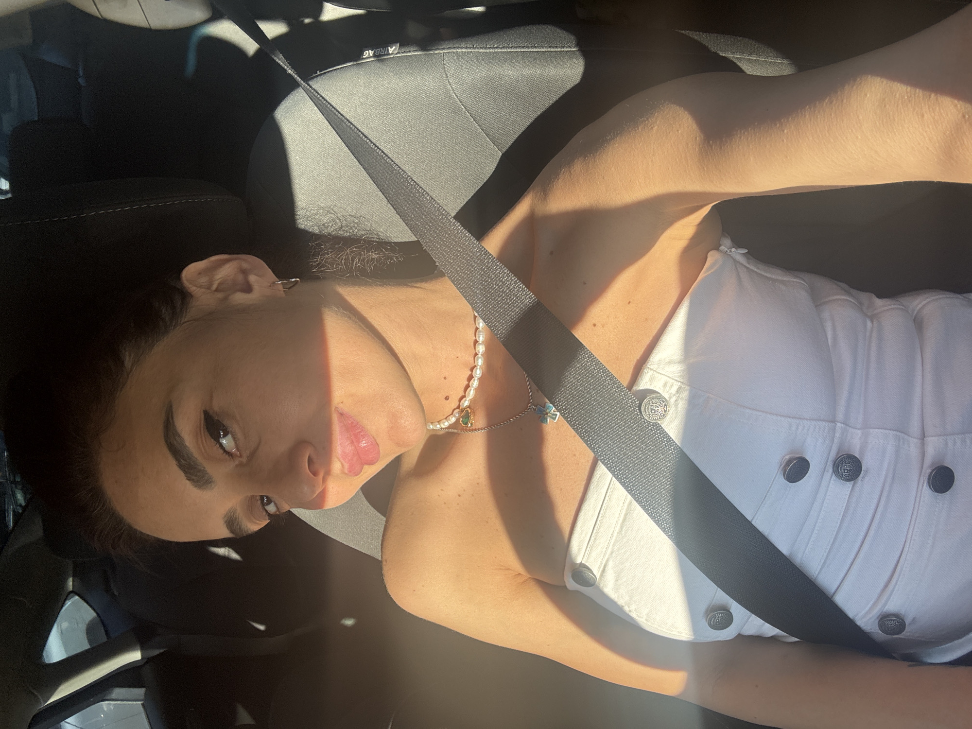 A young woman with dark hair, wearing a white strapless top and a pearl necklace, is seated in the passenger seat of a car. She is looking towards the camera with a slight smile. The sun is shining, casting shadows across her face and shoulder. The interior of the car is visible, including the seatbelt and the headrest. The image was taken in Tbilisi, Georgia, though no specific landmarks are identifiable.