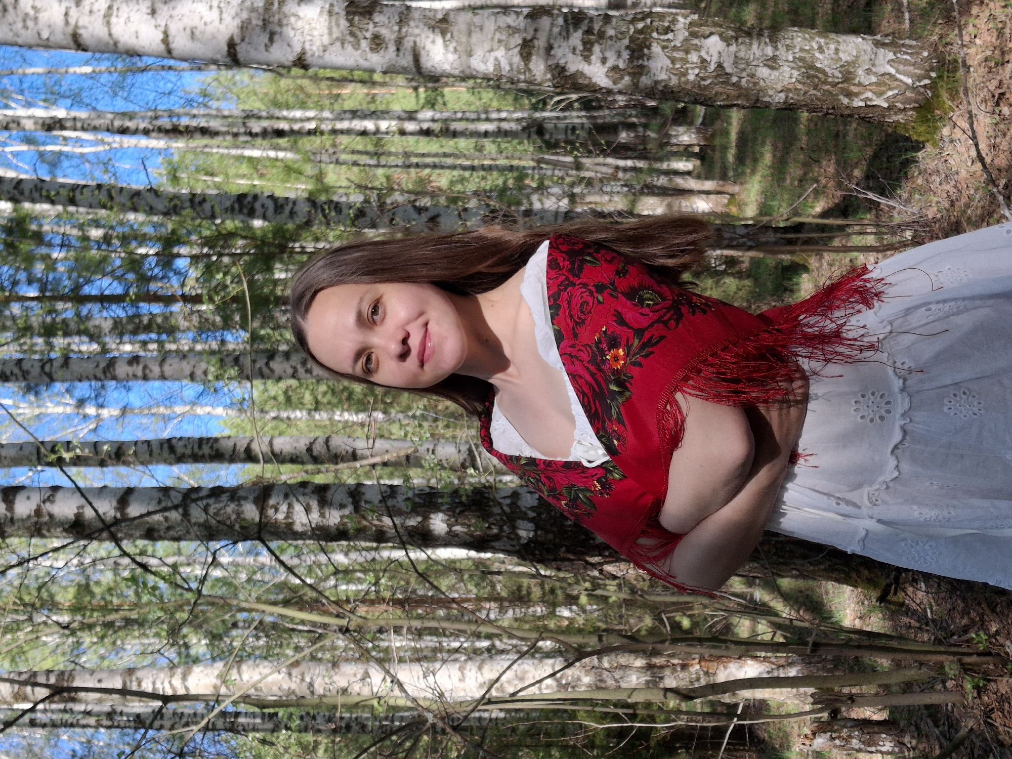 A woman stands in a birch forest, wearing a white dress and a red shawl with fringe. The forest consists of numerous birch trees, with a glimpse of blue sky visible through the canopy. The ground is covered in foliage and small branches.