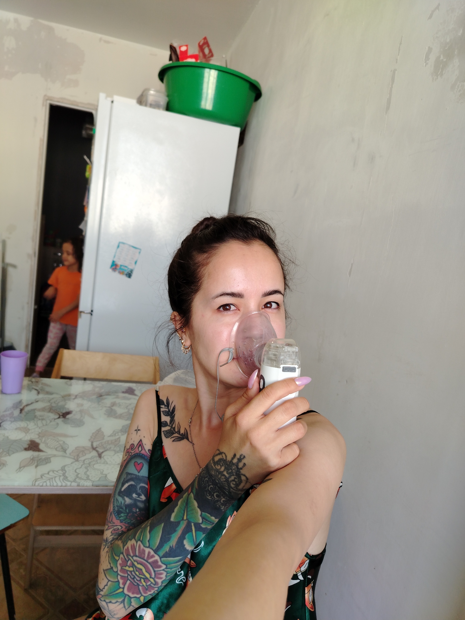 The media file shows a medium shot of a woman with dark hair pulled back in a bun, wearing a green dress and holding a nebulizer mask to her face. She has tattoos on her arms, including a raccoon and flowers. Her fingernails are painted purple. Her expression is neutral, and she is looking at the camera.
In the background, a white refrigerator is visible with a large green plastic tub on top. To the left of the refrigerator is a doorway leading to another room, where a young child in an orange shirt and patterned leggings is walking away from the camera. A table with a patterned tablecloth is in the foreground. The setting appears to be a home interior, likely a kitchen or dining area. The lighting suggests it is daytime. The overall mood is candid and domestic.
There is a word "PUSH" visible on the nebulizer device. No other text is clearly legible. The location context is Noviy, Russia.
