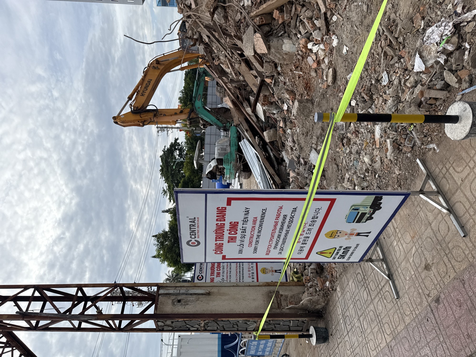 The image shows a construction site in Nha Trang, Vietnam, with demolition in progress. A large pile of rubble, consisting of concrete, bricks, and wood, dominates the right side of the frame. An excavator with the "HYUNDAI" logo is visible, partially obscured by the debris. Another smaller, green excavator is also present.
In the foreground, a temporary construction barrier made of yellow tape is strung between poles. A white sign with red lettering is prominently displayed on a metal stand. The sign contains text in Vietnamese, English, and Russian, stating "CONSTRUCTION AREA SORRY FOR THE INCONVENIENCE" and indicating ongoing construction work. Below this, another sign with a yellow triangle and a construction worker icon warns to "SLOW DOWN TO 5KM/H" and depicts a garbage truck.
To the left of the sign, a tall, rusty metal structure, possibly part of an old building or a utility pole, stands against a cloudy sky. A portion of a white container or structure is also visible behind the metal pole. The ground in front of the sign is paved with patterned tiles. The overall scene conveys a sense of active demolition and construction activity in an urban environment.
