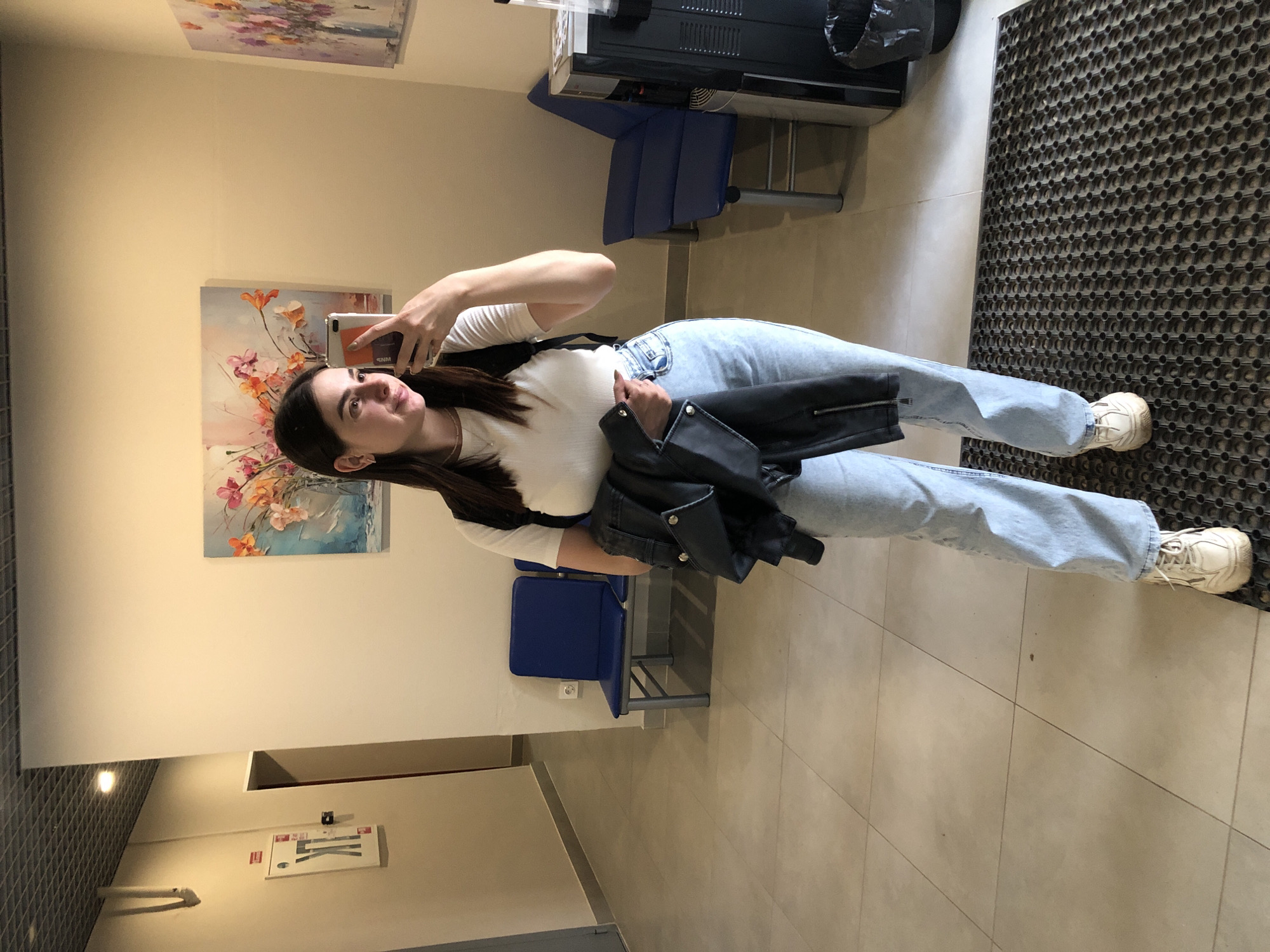 A young woman is standing in what appears to be a clinic or waiting area, taking a mirror selfie with her phone. She is wearing a white t-shirt, light blue jeans, and white sneakers, and is carrying a black jacket over her arm. She has a backpack on. The room has tiled flooring, pale yellow walls, and a few pieces of art on the walls. There are several blue chairs in the background, a water cooler, and a trash can. The image also shows a hallway with a door and some signage on the wall, suggesting it is a public building. The location in Chaikovsky, Russia is not specified in the image.