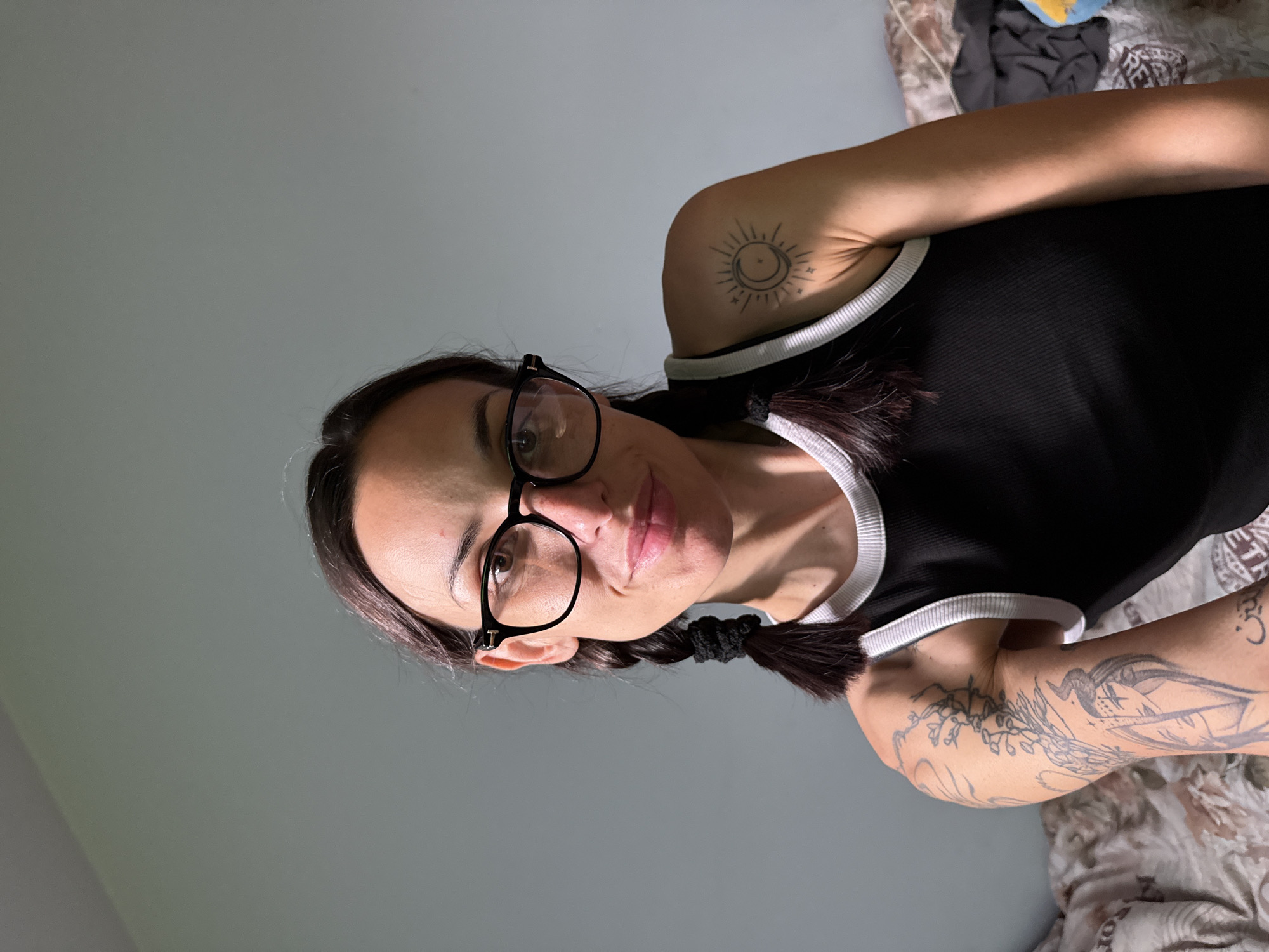 A woman with dark hair styled in pigtails and wearing glasses is pictured from a slightly upward angle against a gray background. She is wearing a black tank top with white trim. The woman has tattoos on her arms and shoulders, one appearing to be a sun and moon design. The lower portion of the image shows a bed covered in patterned bedding, and the top of the frame is plain gray. There is no information available to determine whether the image was taken in Pleven, Bulgaria.