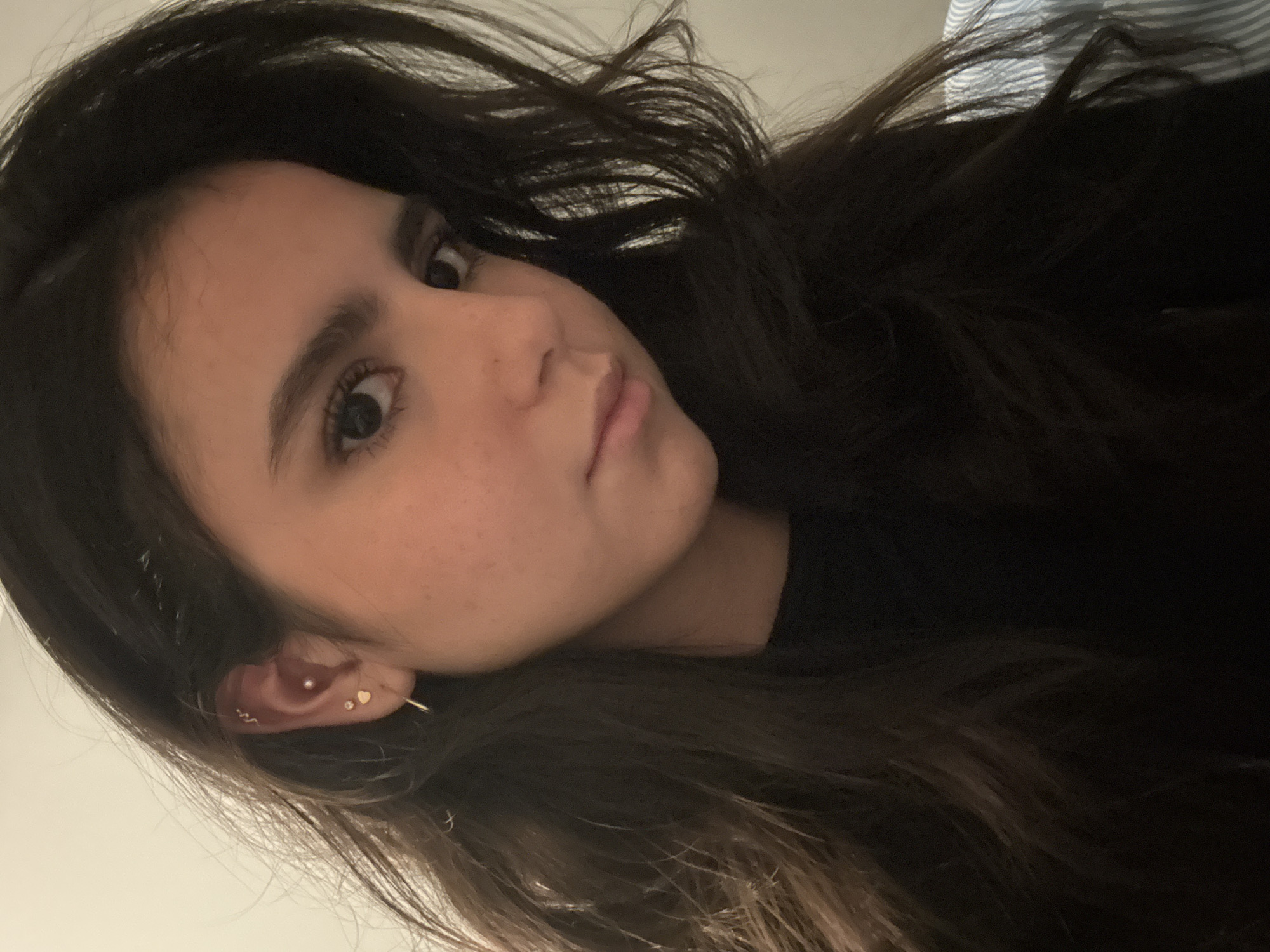 A close-up, tilted portrait features a young person with long, dark brown hair. The subject, appearing to be female, is looking directly towards the camera with a calm and direct gaze. Her head is angled to the right, and her face is positioned in the upper left quadrant of the frame.
Notable features include dark eyebrows, prominent eyelashes, and a subtle, closed-mouth expression. Her skin has a natural appearance with some freckles visible on her cheek. Her left ear is adorned with several small, gold-toned earrings: a hoop or cuff piercing along the upper cartilage, a small stud further down, and two heart-shaped studs in the lobe. A small, shiny gem is also visible in the helix area of the ear.
The subject's dark, wavy hair cascades around her face and shoulder, occupying a significant portion of the background, particularly on the right side of the image. A glimpse of what appears to be a light-colored, possibly blue and white striped fabric is visible behind her hair in the upper right background, suggesting an indoor setting. The lighting is soft and even, highlighting the subject's features. No text is visible in the image.