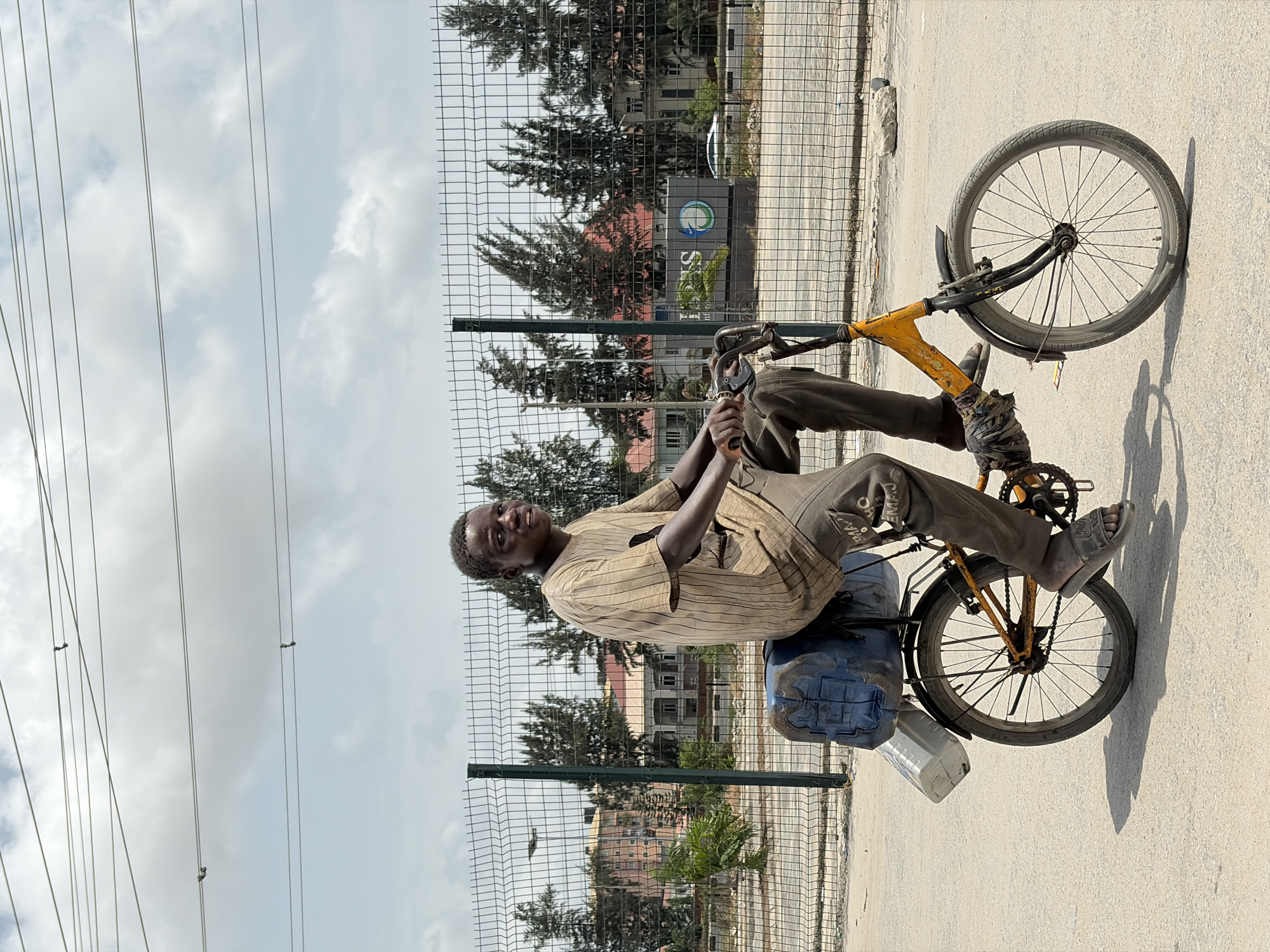 A man is sitting sideways on a bicycle in Igboefon, Nigeria. The man is of African descent and is wearing a striped shirt and pants. The bicycle is heavily used, with a yellow frame and black tires. The man has some sort of containers behind him on the bicycle. He is sitting on these containers. A metal fence is visible behind the man, and beyond that, trees, and buildings. Power lines run across the top of the image. The ground is paved.