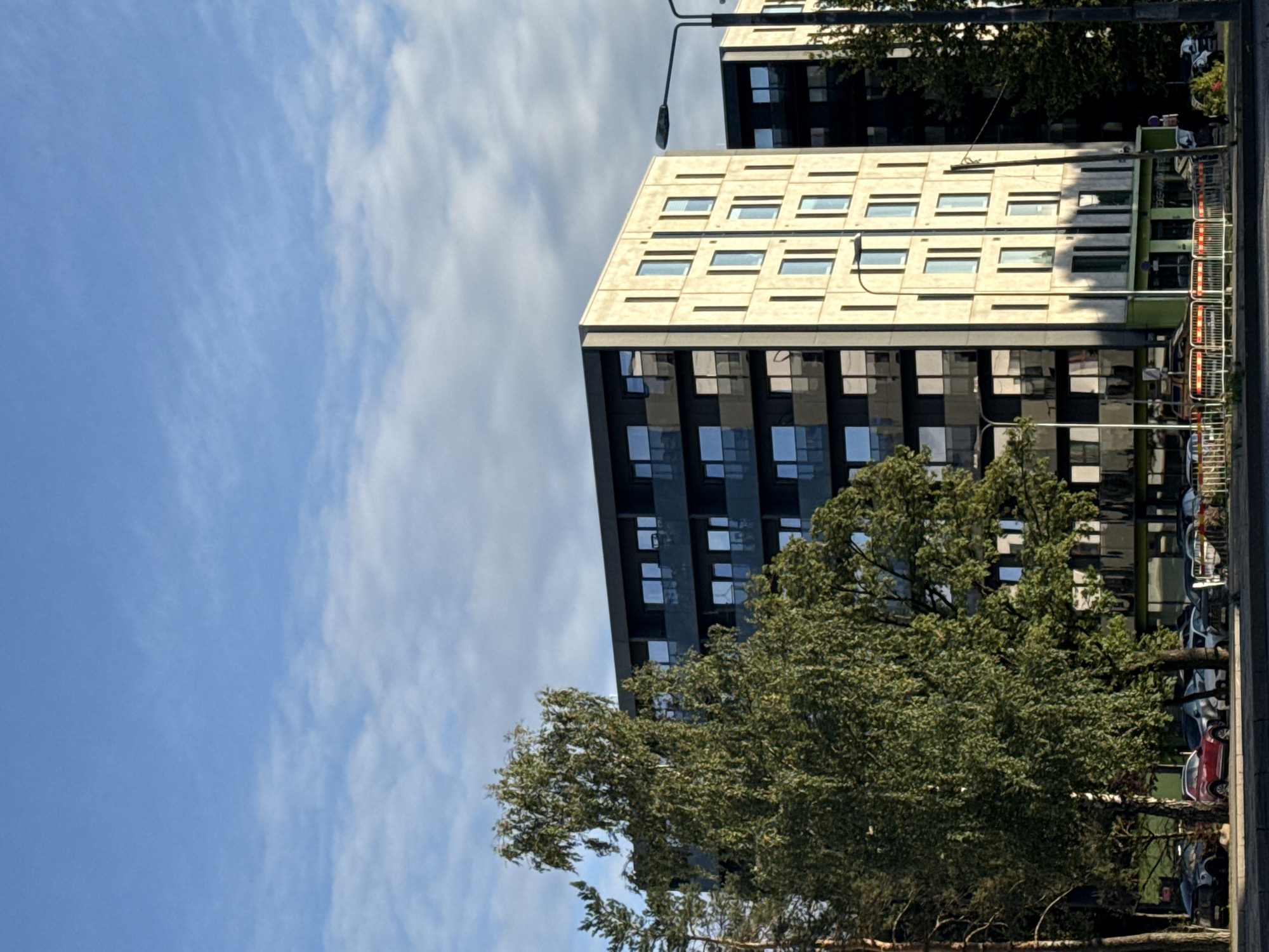 A vertical, medium shot captures two modern apartment buildings in Tallinn, Estonia, on a partly cloudy day. The building on the left, in the foreground, is characterized by a pale concrete facade with vertical window sections and dark, recessed balconies. The building to its right is darker, appearing to have a black facade with large, reflective windows.
In the lower left quadrant, a large deciduous tree with light green, drooping leaves partially obscures the left building. To the left of this tree, a pine tree is visible. The ground level of the left building features a green-colored section with windows, suggesting commercial spaces.
The street in front of the buildings is lined with parked cars, including a red SUV, a dark hatchback, and several other vehicles. Temporary white and orange barriers are positioned along the sidewalk. A lamppost with a dark fixture extends from the right side of the frame, casting a shadow on the pale facade of the building. The sky above is a vibrant blue, with scattered white clouds.