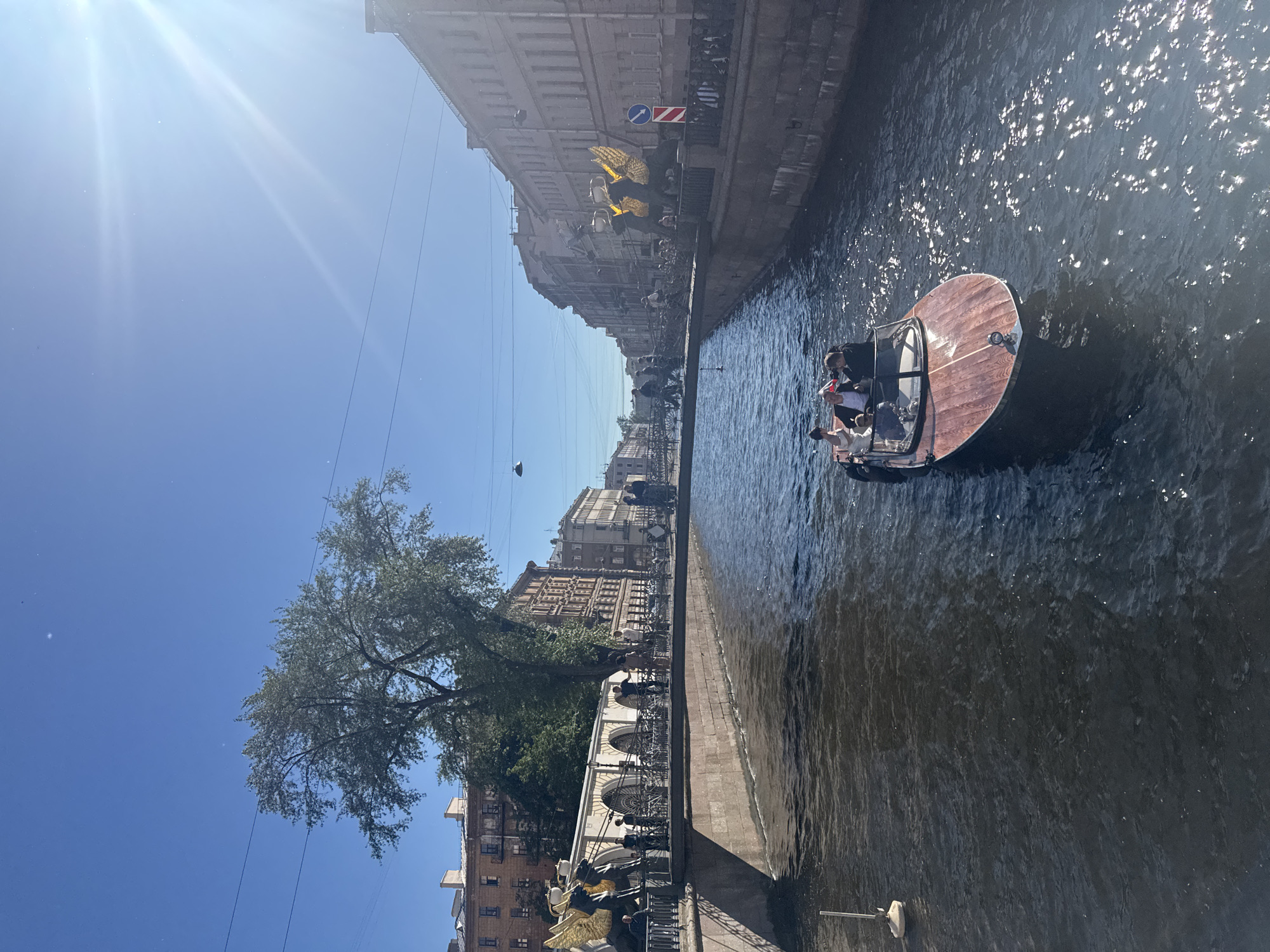 This media file captures a sunny day scene along a canal in Saint Petersburg, Russia. The main subjects are a small, open-topped wooden boat with several people aboard, navigating the dark, rippling water. The boat has a polished wooden deck and a partial canopy. The passengers appear relaxed, enjoying the ride.
The canal is lined with historic buildings, characterized by classical architecture with multiple stories and ornate facades. One building prominently features a row of arched windows. Along the canal's edge, a stone embankment is visible, bordered by a decorative metal railing. Trees with lush green foliage overhang the water, casting shadows. In the distance, more buildings and a bridge can be seen.
The bright sunlight suggests it's midday. The sky is a clear, vibrant blue with some lens flare in the upper left corner, indicating the sun's intensity. The overall atmosphere is peaceful and leisurely, with people enjoying a boat tour on a warm day. There are no visible text elements in the image.