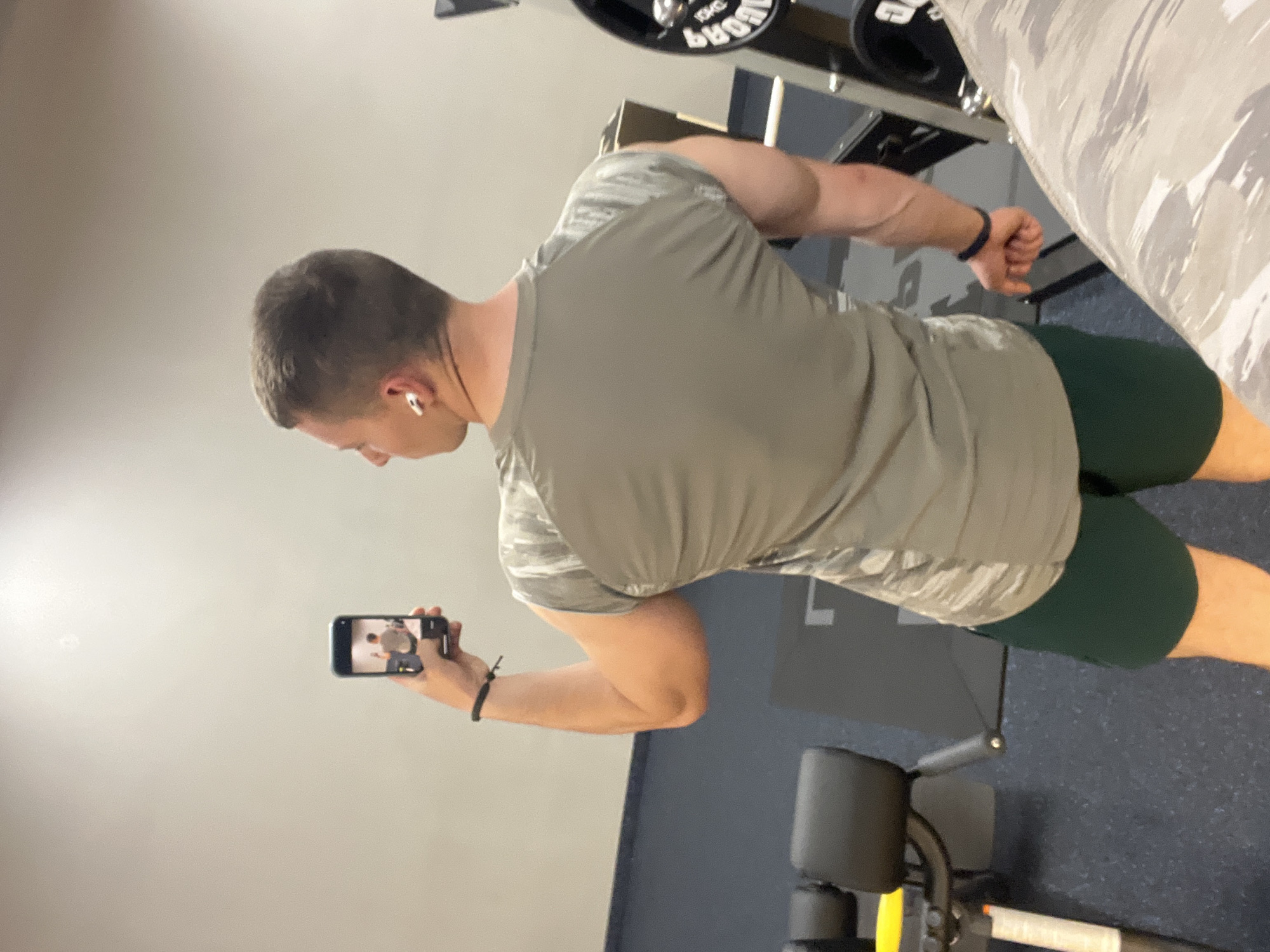 A man is in a gym setting taking a selfie with his cell phone. He is wearing a green T-shirt and green shorts. He has earbuds in his ears and a black wristband on his wrist. The gym has workout machines and a weight rack with weights on it. The floor is a blue-gray color. There is a light-colored wall behind him. The specific location in the city of Burgas, Bulgaria, cannot be determined from the image.