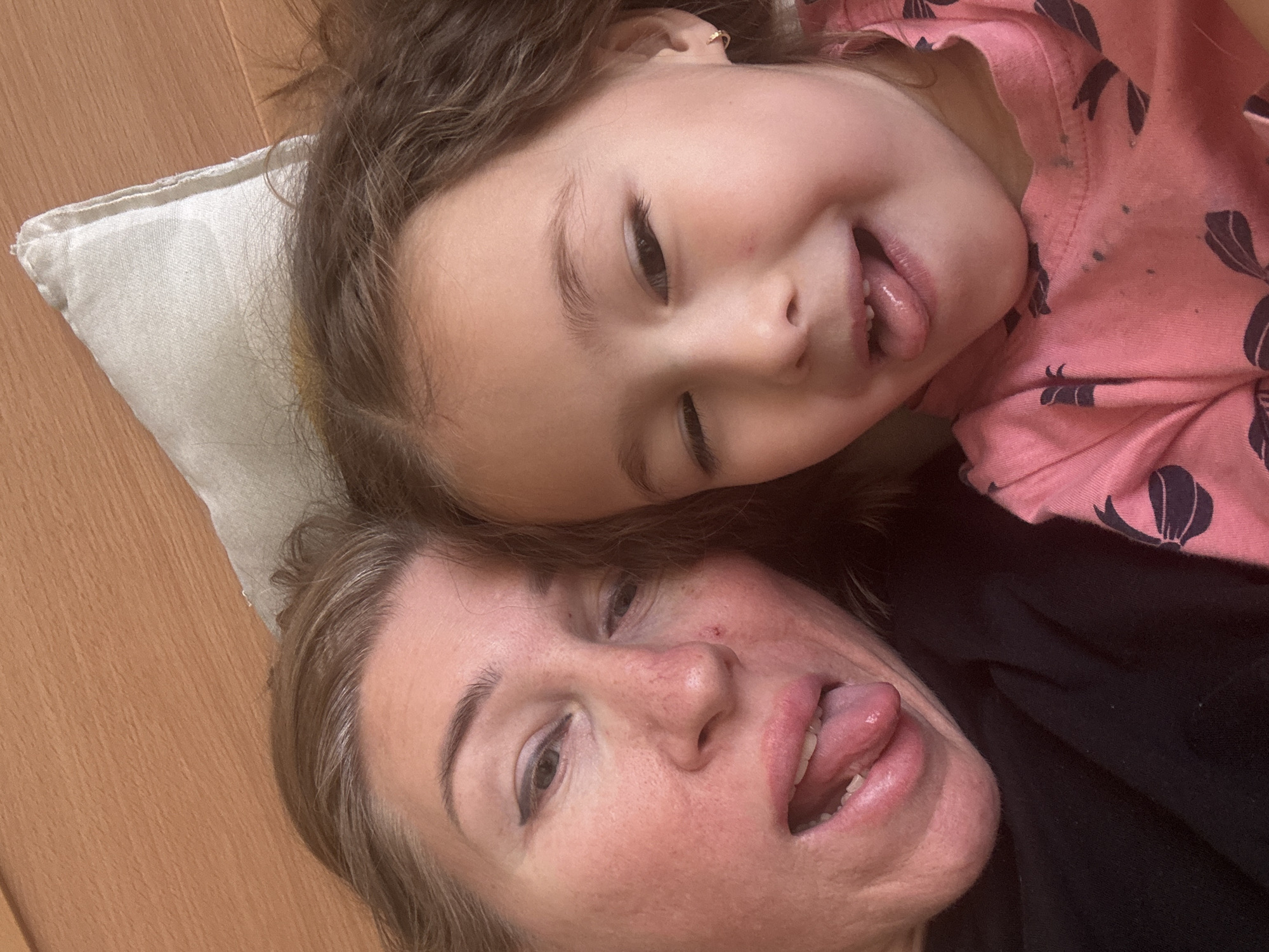 A selfie-style close-up shot depicts a woman and a young girl lying down together, both sticking out their tongues. The woman has blonde hair and is wearing a black top. The girl, who appears to be her daughter, has brown hair and is wearing a pink shirt with a dark floral pattern. They are lying on a light-colored pillow on a wooden floor. The location is unspecified, and there is no evidence to suggest it is in Tashkent, Uzbekistan.