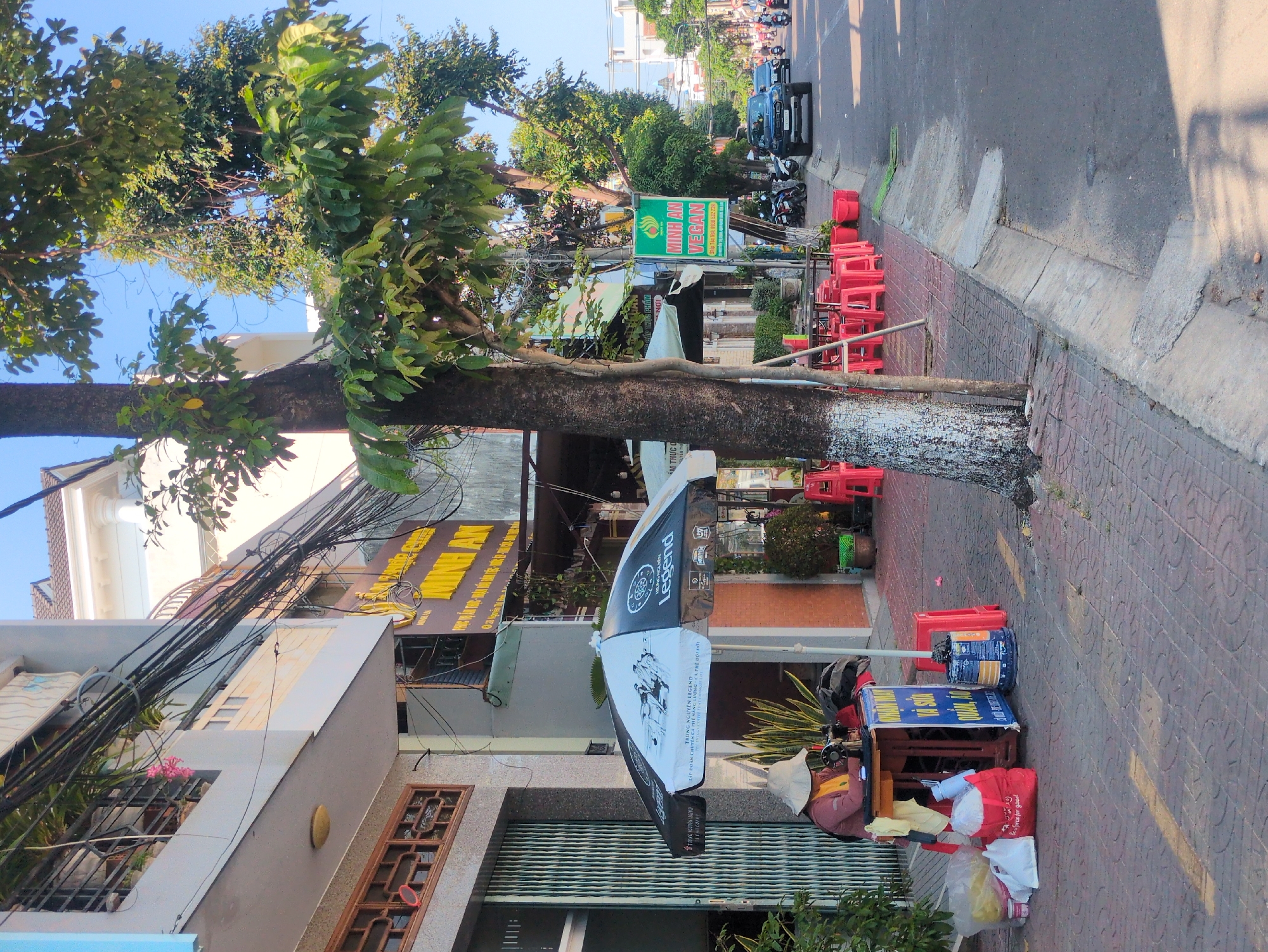 The image shows a street scene in South Quy Nhon Ward, Vietnam. On the right side, there is a tiled sidewalk with red chairs lining the edge and a tree with a painted lower trunk. Adjacent to the sidewalk, there is an asphalt street with vehicles and motorcycles parked on the side. A sign reads "MINH AN VEGAN". On the left, there is a multi-story building with a sign that reads "ẨM THỰC". An umbrella with a "Legend" logo is partially visible. Near the building, a street vendor is operating with various items including bags, a sewing machine, and a blue sign with text. Above the building, there are numerous entangled power lines. The sky is clear and blue.