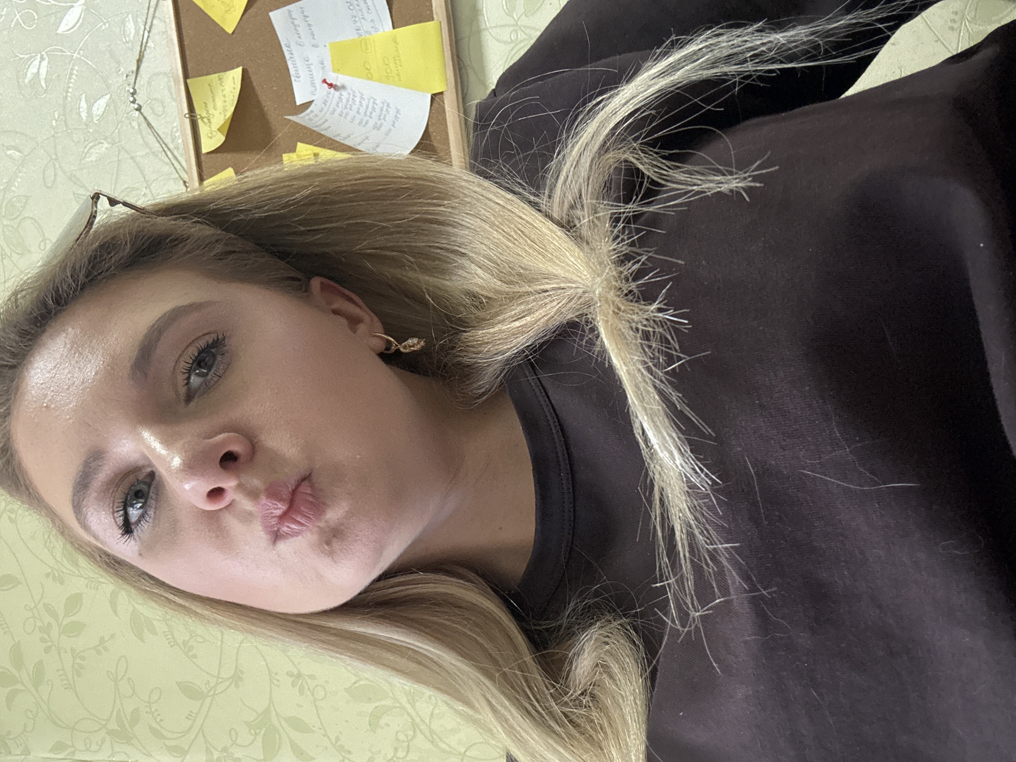 A blonde woman with blue eyes and light skin is taking a selfie. She is wearing a dark brown crew neck shirt and a gold hoop earring in her left ear. She is puckering her lips and looking at the camera. Behind her, a bulletin board with yellow sticky notes is visible. The wall behind the bulletin board has a light green floral pattern. Her hair is long and blonde, and some strands are loose around her face and chest. She appears to be indoors, possibly in an office or home environment.