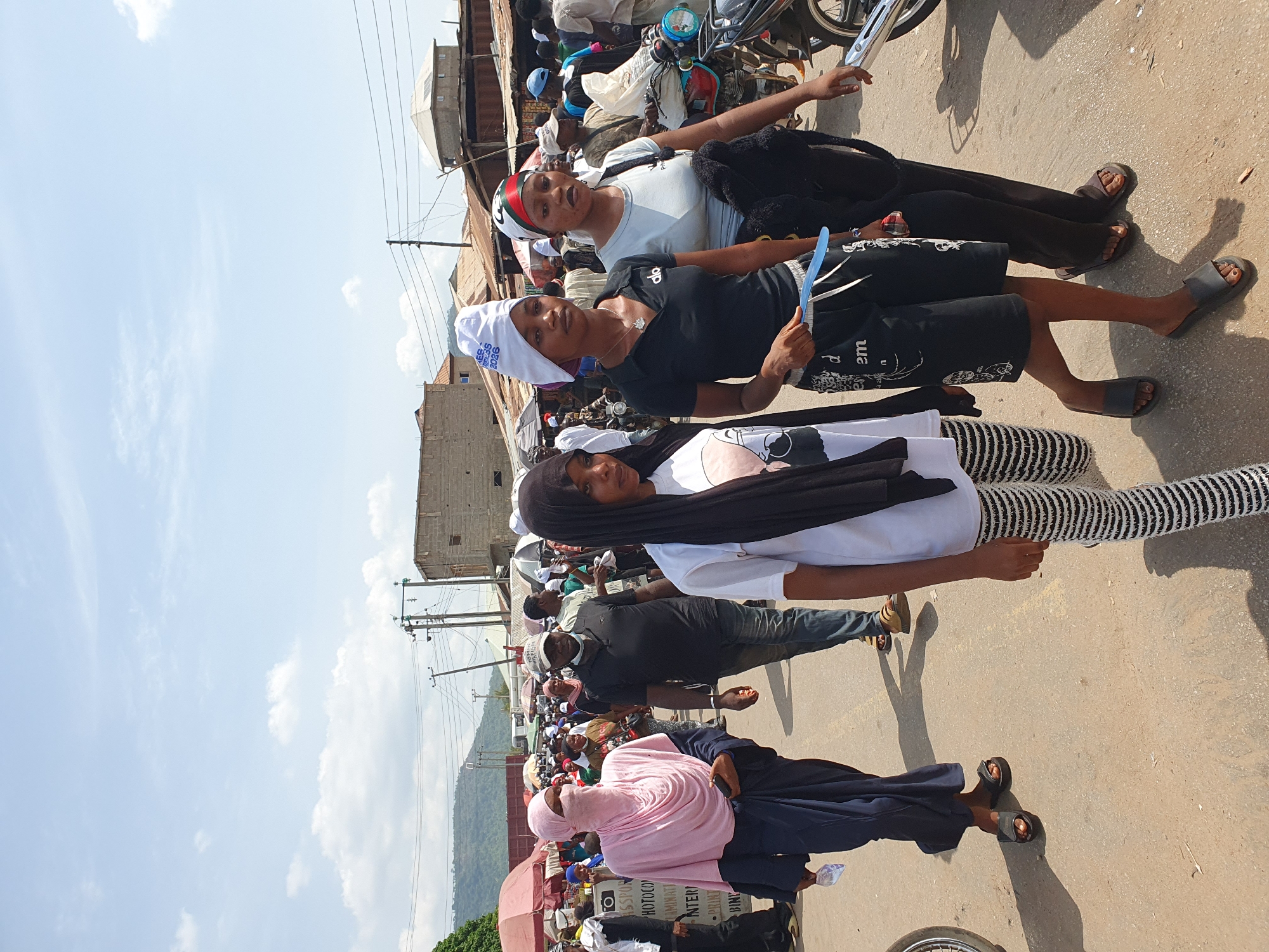 The image depicts a street scene in Lokoja, Nigeria, under a partly cloudy sky. A group of people are walking or standing on a paved road. The individuals are of varying ages and are dressed in diverse styles of clothing. Some of the people are wearing head coverings. There is a mix of casual wear, traditional attire, and religious garb. In the background, there are buildings and structures of varying heights and materials, indicative of an urban environment. Overhead electrical wires crisscross the sky. A small hill or mountain can be seen in the distance. Motorbikes are also present.
