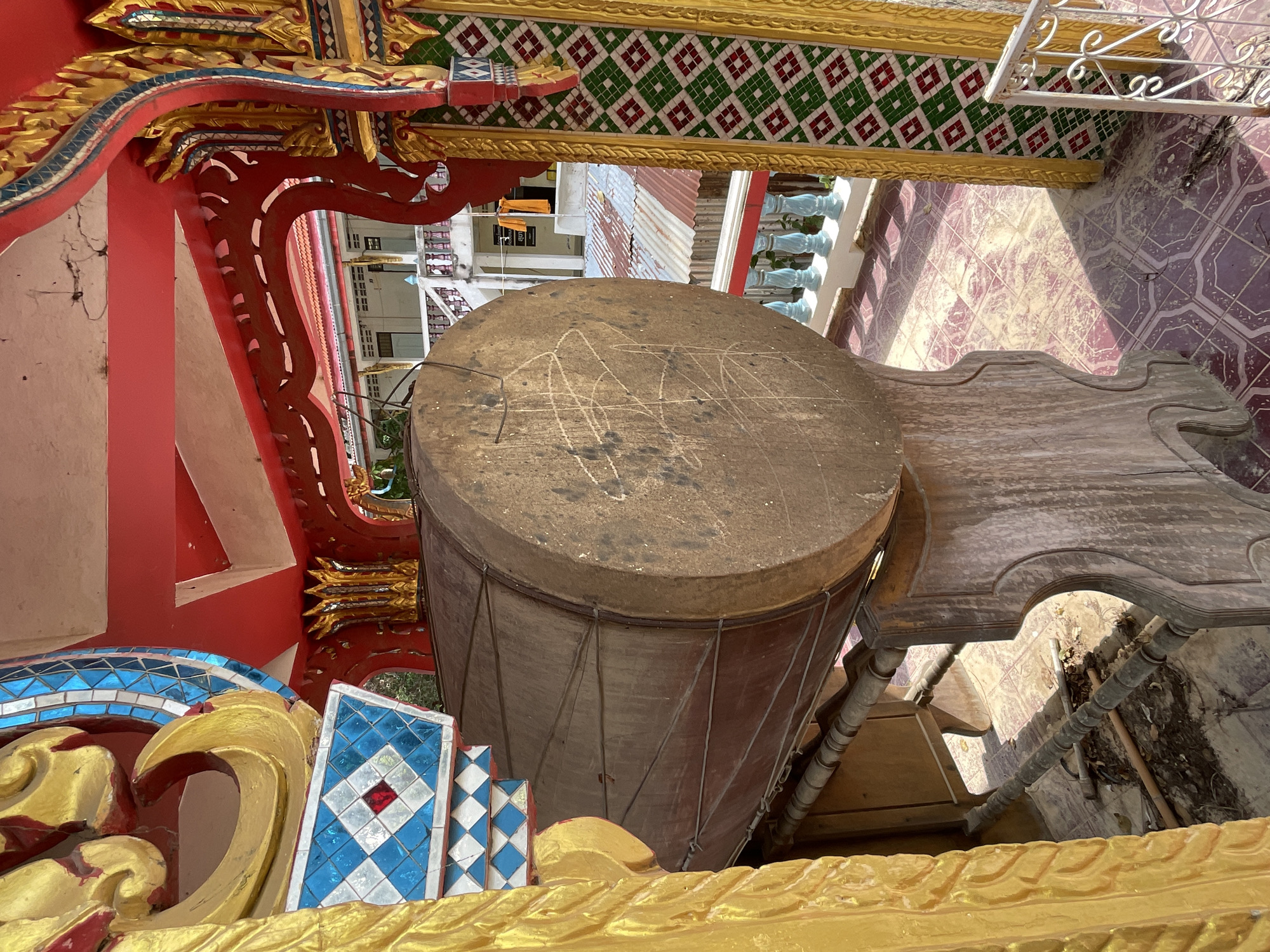 An outdoor medium shot captures a large, weathered drum resting on a wooden stand within a covered structure. The drum, predominantly brown and showing signs of age and use with visible scratches on its surface, is positioned to the left of the frame. It is supported by a dark, curved wooden stand. The background reveals architectural elements of a building, possibly a temple, with vibrant red and gold ornate decorations. Blue and white tiled patterns are visible on the left side, adjacent to the drum. Further back, a building with white walls and a series of windows can be seen, along with a metal railing and a corrugated metal roof. The ground is tiled with a pattern of pink and purple squares. The scene suggests an outdoor setting in Pattaya City, Thailand, likely part of a cultural or religious site.