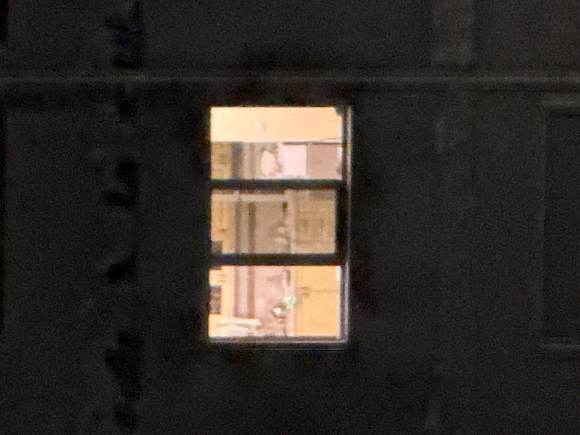 The image shows a dark, grainy exterior view of a building at night, with a brightly lit rectangular window as the central focal point. The window is divided into at least three panes. Through the window, the interior of what appears to be a kitchen is visible. Yellowish light emanates from the interior, illuminating wooden cabinets and countertops. Several objects are present on the countertops, though their details are indistinct due to the distance and image quality. The overall scene suggests a residential building in Rafailovići, Montenegro, viewed from the outside after dark, with the light from the kitchen window providing the only significant illumination in the composition. The surrounding building facade is dark and indistinct, with some vague shapes or textures visible in the upper portion of the frame.