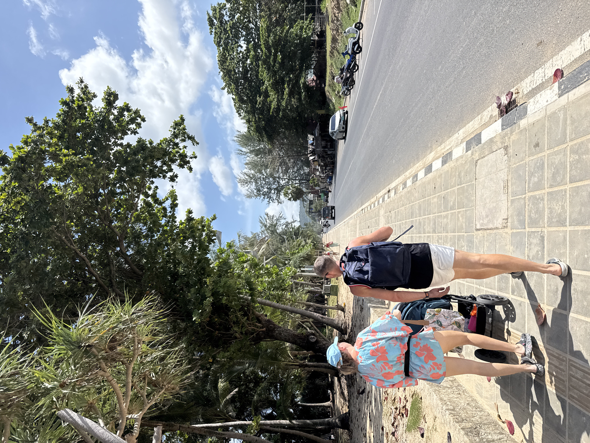 In Karon, Thailand, a man and a woman walk side-by-side on a paved sidewalk next to a busy road. The woman, wearing a blue and pink floral shirt and a blue cap, pushes a stroller. The man, with a dark blue backpack, wears white shorts and a black tank top. The sidewalk is bordered by sand and lush green trees, including palm trees. Across the road, shops and parked motorcycles are visible. The sky is a bright blue with scattered white clouds, suggesting a sunny day.