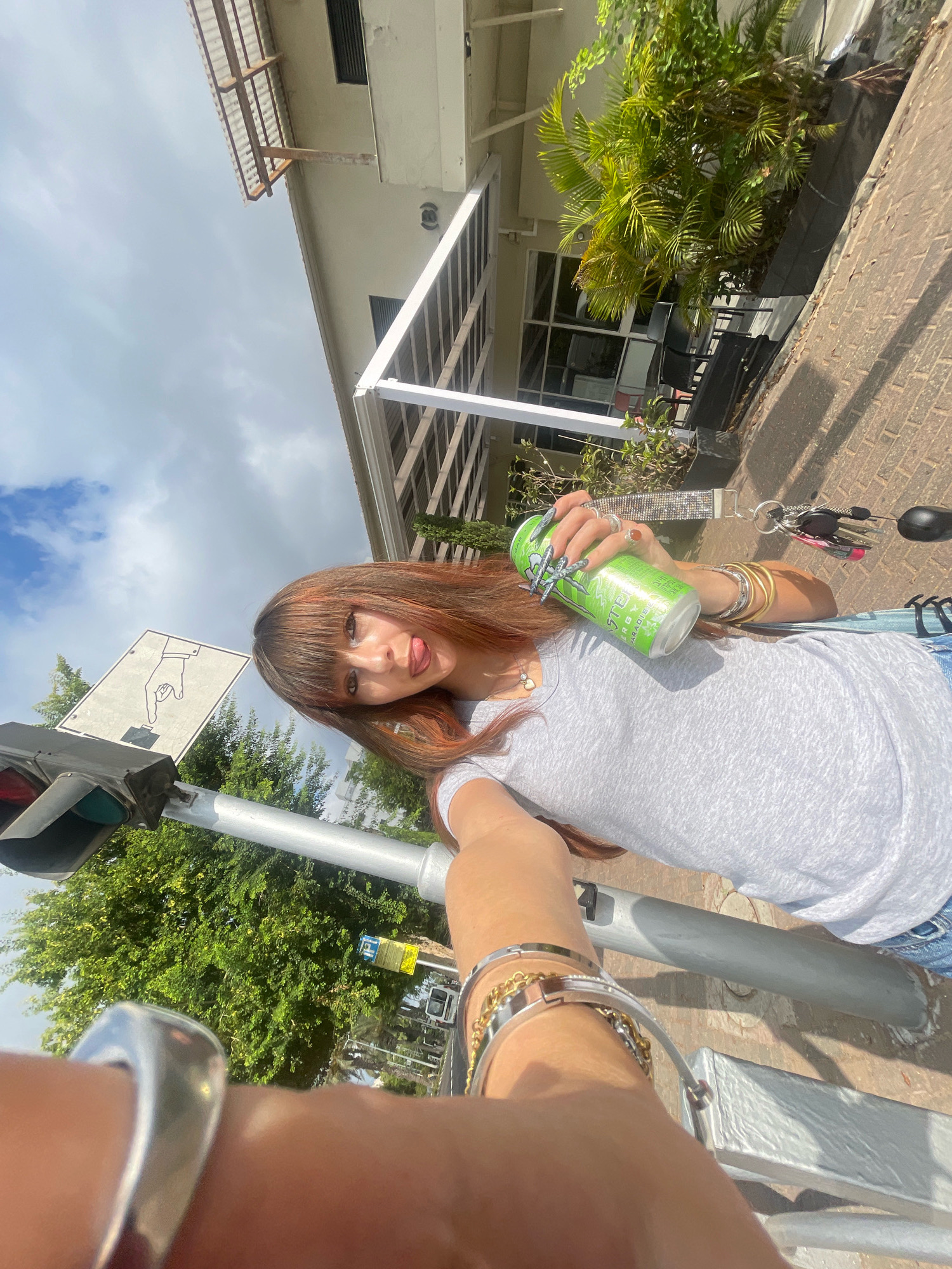 A woman with auburn hair and bangs is standing on a sidewalk in Raanana, Israel, holding a green energy drink can and looking towards the camera. She is wearing a grey t-shirt and blue jeans. The background shows a building with a white awning, palm trees, and a traffic light. The image is taken from a slightly low angle, with the sky and clouds visible above.