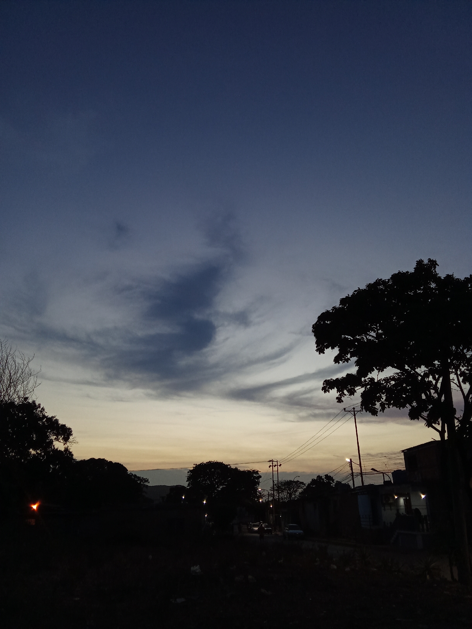 The image shows a dusk or dawn scene in what appears to be a residential area in Juan Griego, Venezuela. The sky is a gradient of dark blue to lighter shades of grey and yellow near the horizon, with some distinct cloud formations. The foreground is mostly dark, with silhouetted trees and foliage. Further back, there are signs of a settlement with houses and utility poles. There are street lights visible. Vehicles can be seen parked on the side of the road. The overall scene conveys a sense of stillness and quietness.