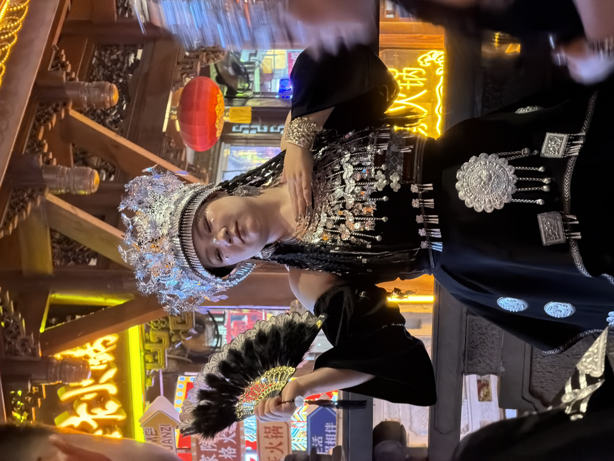 A woman in traditional attire, adorned with a silver headdress and ornate jewelry, holds a black feather fan while standing in what appears to be a street market in Yuzhong District, China. The background features glowing neon signs in Chinese characters, traditional wooden architecture, and red lanterns, suggesting a lively cultural setting. The woman's outfit is primarily black, detailed with intricate silver embroidery and dangling ornaments. Her pose and expression suggest a cultural performance or demonstration.