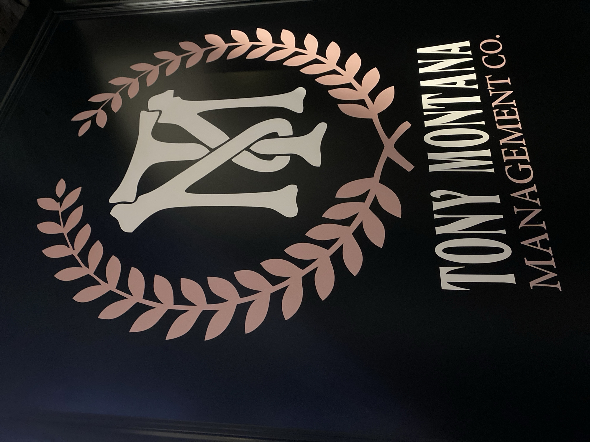 The image displays a professionally designed sign or door, predominantly black, featuring a stylized logo and text. The scene is static, showcasing the branding of a company.
The logo consists of intertwined, ornate letters, likely "T" and "M," presented in a light beige or cream color. These letters are encircled by a laurel wreath design, rendered in a distinctive rose gold or copper hue. The overall aesthetic of the logo suggests a blend of classic and modern, with its serif-like letterforms and traditional wreath motif.
To the right of the logo, the company name "TONY MONTANA MANAGEMENT CO." is displayed vertically. "TONY MONTANA" is written in the same light beige/cream serif font as the intertwined letters of the logo. Below this, "MANAGEMENT CO." is rendered in the matching rose gold/copper color of the laurel wreath, maintaining a consistent brand identity.
The background is a smooth, dark surface, possibly a door or a wall sign, against which the lighter-colored graphics stand out sharply. There are subtle reflections on the black surface, particularly towards the bottom, suggesting a polished finish and indoor lighting. The image is taken from a slight upward angle, capturing the full extent of the sign.
No people or explicit activities are visible, focusing solely on the brand's presentation. The clean lines and clear text indicate a professional setting. The location context specifies Petrich, Bulgaria.