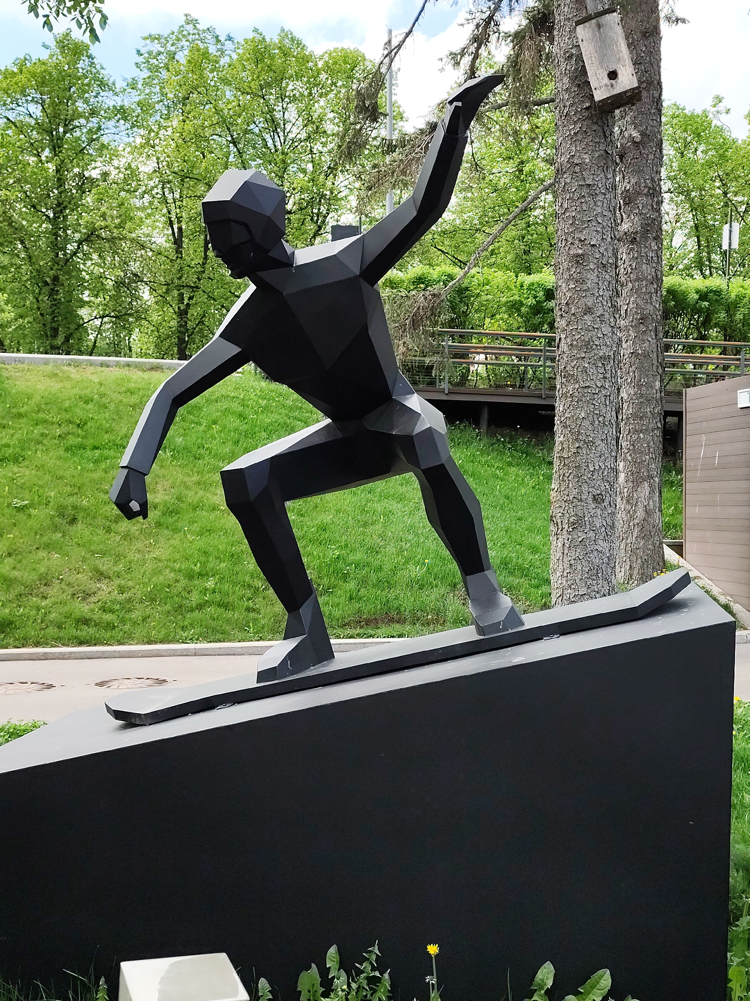 Here's a detailed description of the image:
The image shows a black, geometric, cubist-style sculpture of a snowboarder. The sculpture is mounted on a black pedestal, positioned outdoors in a grassy area.
Behind the sculpture, there's a lush green landscape, including grassy slopes and leafy trees, suggesting a park or recreational area. To the right of the sculpture, a tree trunk is visible, with a wooden birdhouse attached to it. A wooden boardwalk or raised pathway with metal railings can be seen in the background.
The scene is brightly lit, indicating it is daytime, and the presence of shadows gives depth to the image. The overall setting suggests a well-maintained urban park or public space.
Based on visual evidence, including the landscaping and the sculpture's style, this photograph may be from Gorky Park in Moscow, Russia.