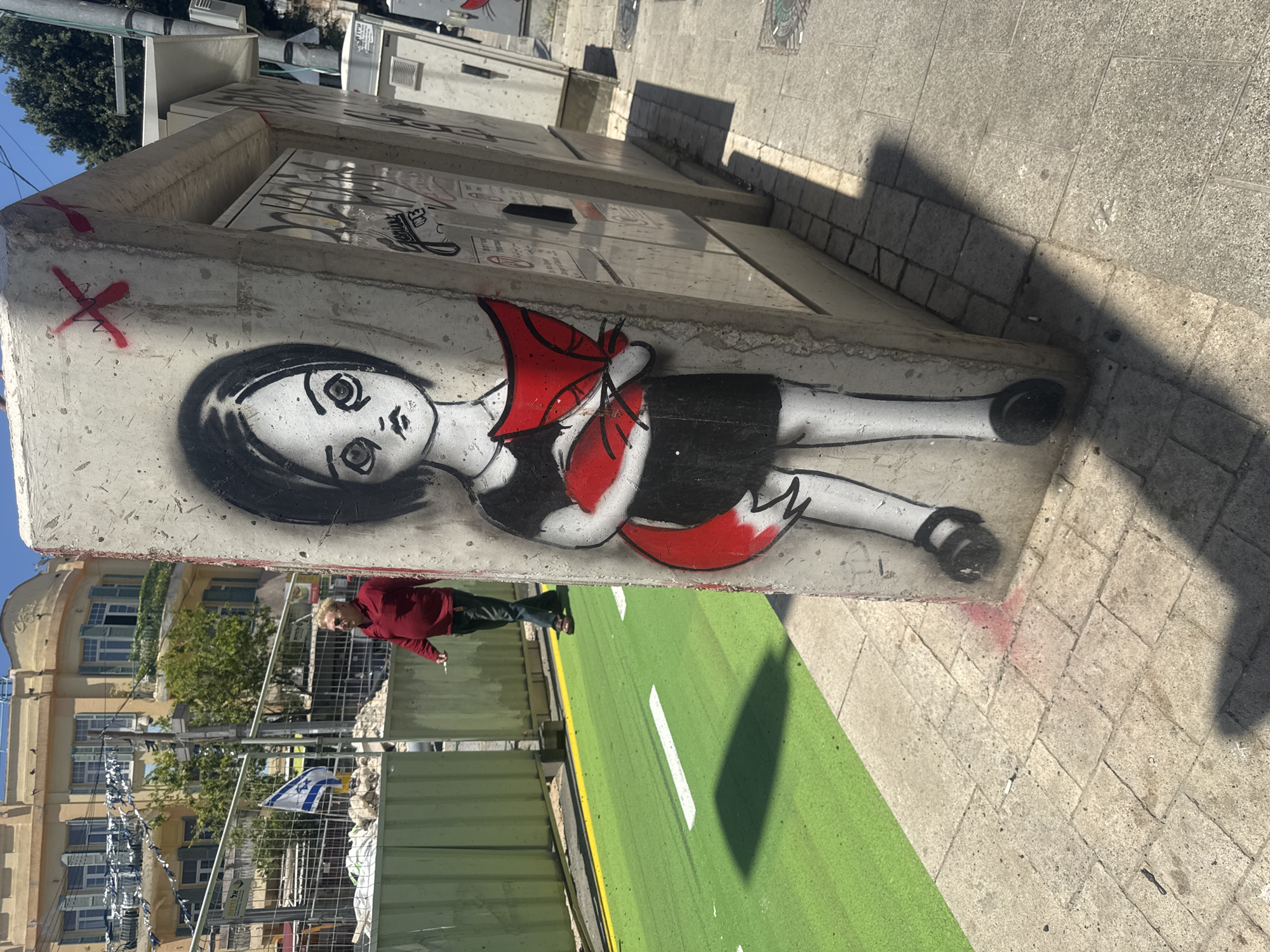 The image shows a graffiti artwork of a young girl with large eyes and a red bow, painted on a concrete utility box in Tel Aviv, Israel. The graffiti is located next to a green-colored bike lane with a single white dividing line. In the background, a building with light-colored walls, green shutters, and trees is visible. A construction fence with the Israeli flag is also in the background. A woman in a red shirt is standing next to the utility box, smoking a cigarette. The ground is paved with light-colored stones. Other utility boxes with graffiti are visible in the background. The lighting suggests it's daytime.