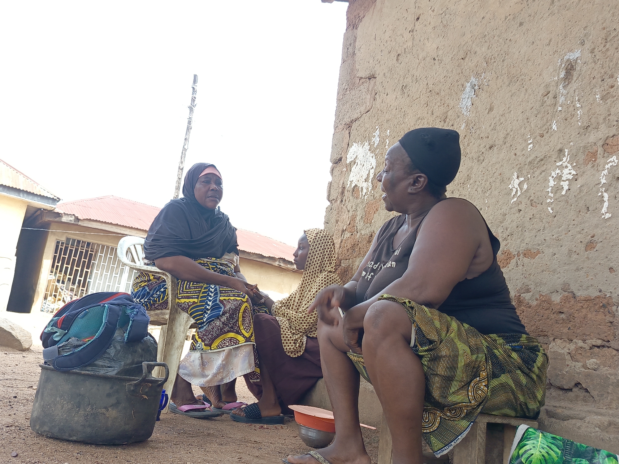 Three women are sitting outside near a building in Akwanga, Nigeria. The woman on the left is seated on a white plastic chair, wearing a dark top, a headscarf, and a colorful patterned skirt with a white slip underneath. Her feet are clad in pink sandals. Beside her is a large, black metal pot with a dark bag and a backpack placed on top. The woman in the middle wears a yellow and brown patterned headscarf and a maroon dress. The woman on the right is sitting on a small wooden stool, wearing a black tank top and a patterned skirt. She has a black cap on her head and is gesturing with her right hand. The building behind them has a rough, textured wall, and the ground is dirt. In the background is a building with a red roof.