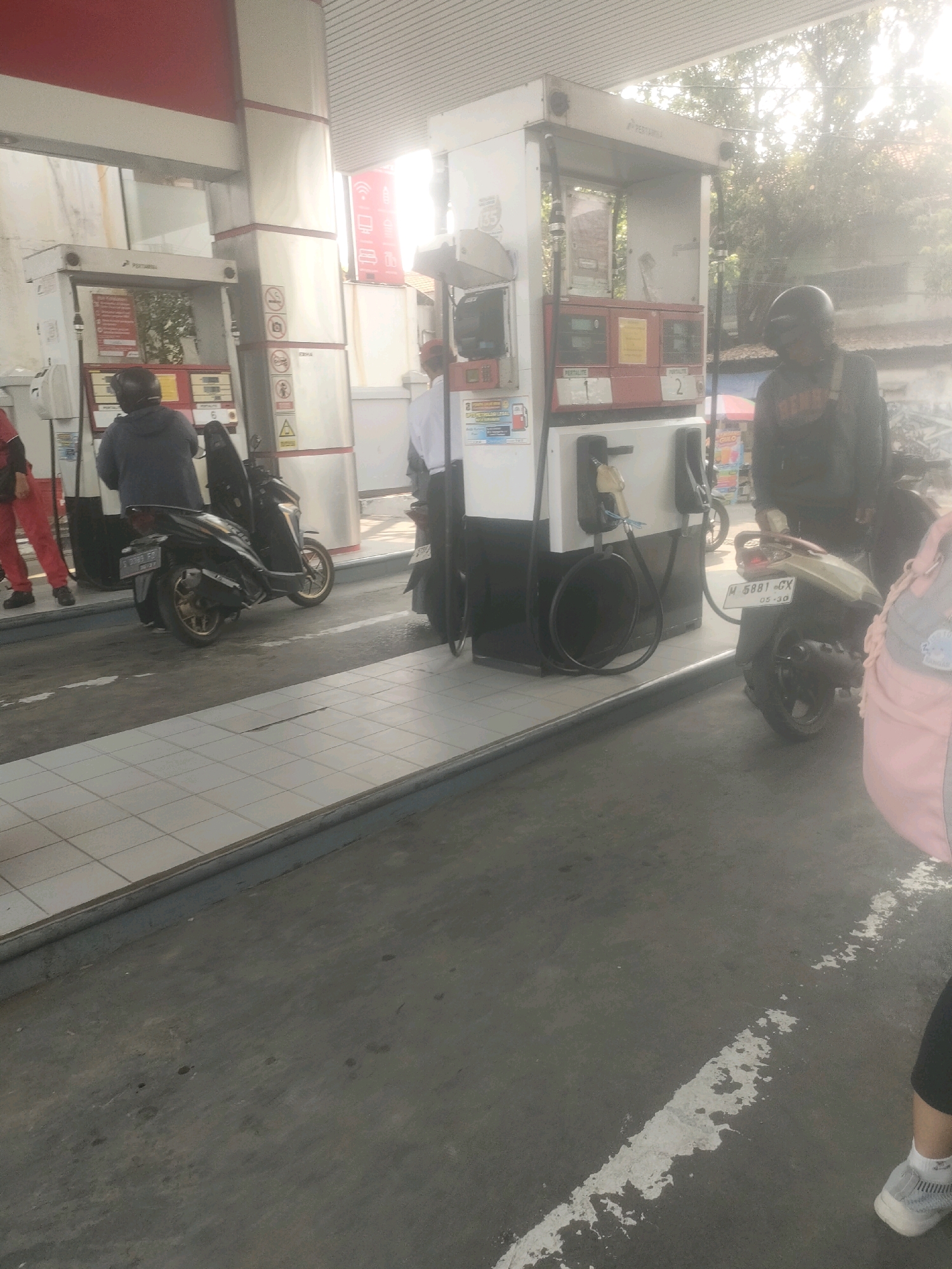 The image depicts a gasoline station in Surabaya, Indonesia. The foreground is dominated by a grey asphalt ground with a white painted line. In the middle ground, a row of gas pumps stands, with a tiled walkway in front of them. Several people are present, some on motorcycles. On the left, a person in red pants stands near a gas pump. A person in a dark jacket is seated on a dark-colored motorcycle. Further to the right, another person in a dark jacket and helmet stands beside a lighter-colored motorcycle. The background shows trees and buildings, suggesting an outdoor urban environment. The gas pumps appear to be from the Pertamina brand, a common state-owned oil and natural gas company in Indonesia. The lighting suggests it is daytime, possibly with some atmospheric haze or glare.