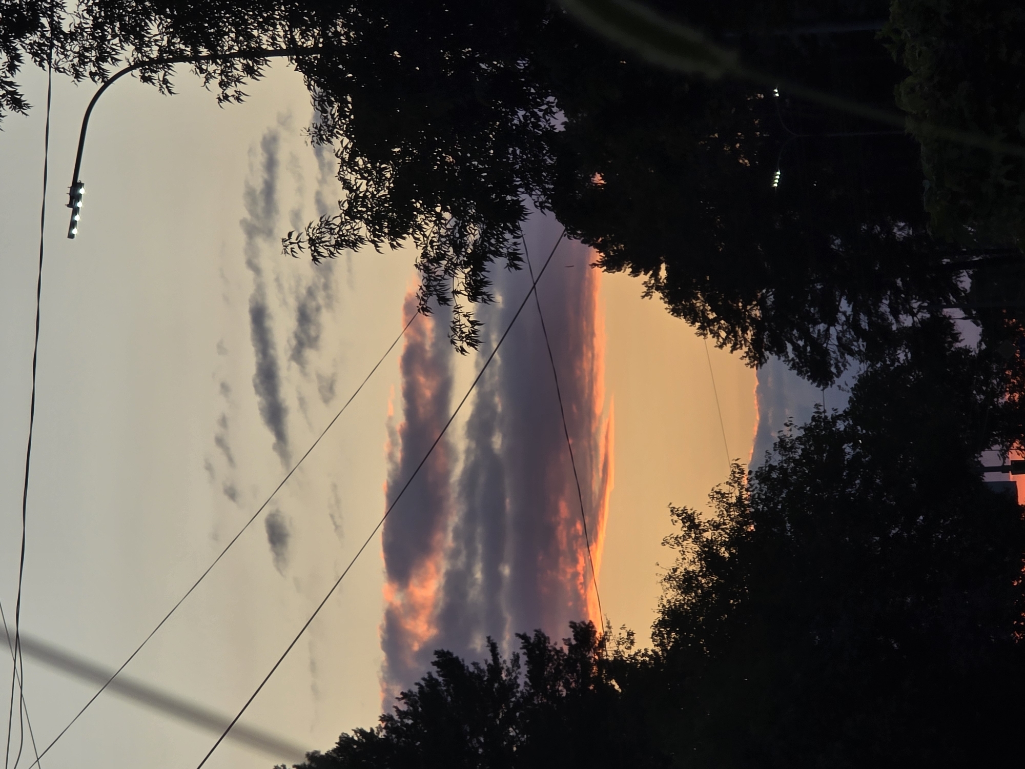 The image captures a sunset over the city of Chaikovsky, Russia. Silhouetted trees frame the view of a sky painted with hues of orange, pink, and purple, with scattered clouds. Power lines crisscross the sky, and a street lamp with four illuminated bulbs is visible on the right side of the frame. The overall scene depicts a serene evening in an urban environment.