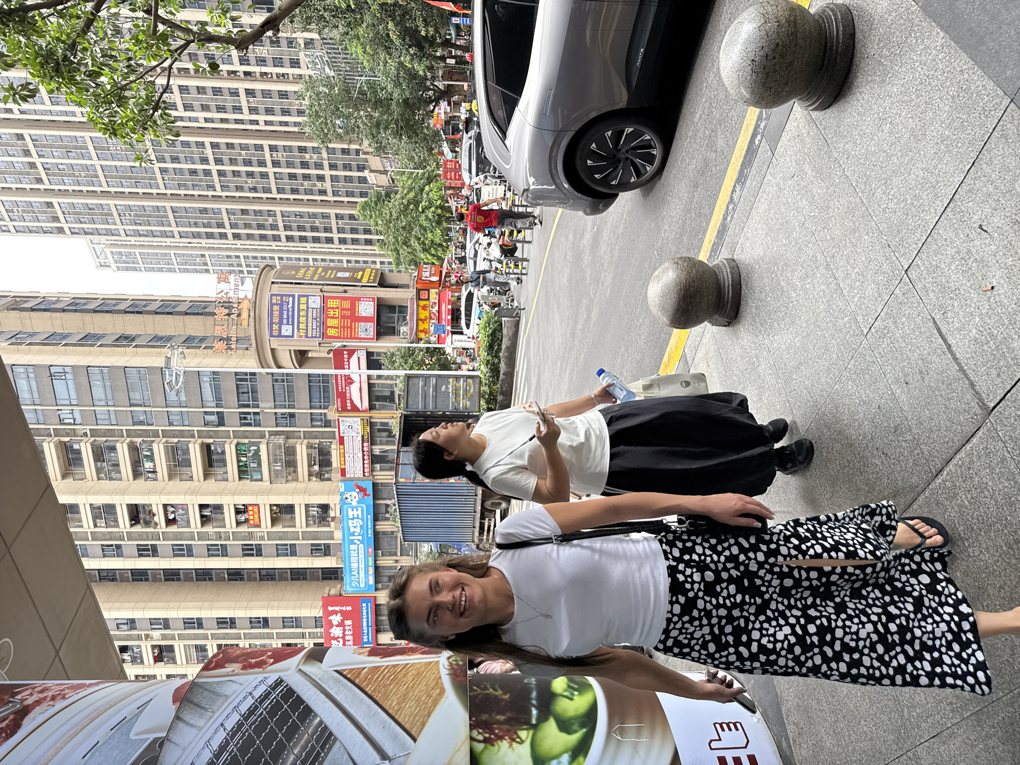 Two women are walking on a sidewalk in Haizhu District, China. The woman in the foreground, with blonde hair, is wearing a white t-shirt, a black and white patterned skirt, and flip-flops. She is smiling and holding a phone in her left hand. The woman behind her, with dark hair, is wearing a white t-shirt and a black skirt. She is holding a water bottle and a phone. They are walking past a building with many windows and balconies, and a large advertisement on the left. In the background, there are tall apartment buildings, a truck, cars, and people walking on the street. The sidewalk is paved with grey tiles and there are two round stone bollards. The scene appears to be a commercial or residential area in a city.