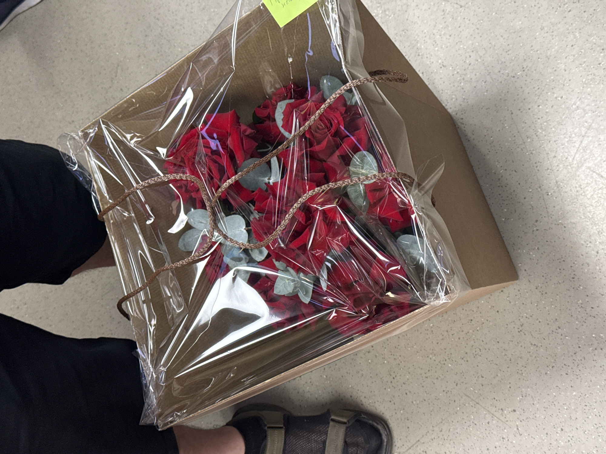 A person is holding a box of red roses wrapped in clear plastic. The box is secured with a brown braided cord. A small yellow sticky note is attached to the box. The person is wearing black pants and dark-colored sandals. The background is a light-colored speckled floor. There is no evidence to suggest that the location is in Moscow, Russia.