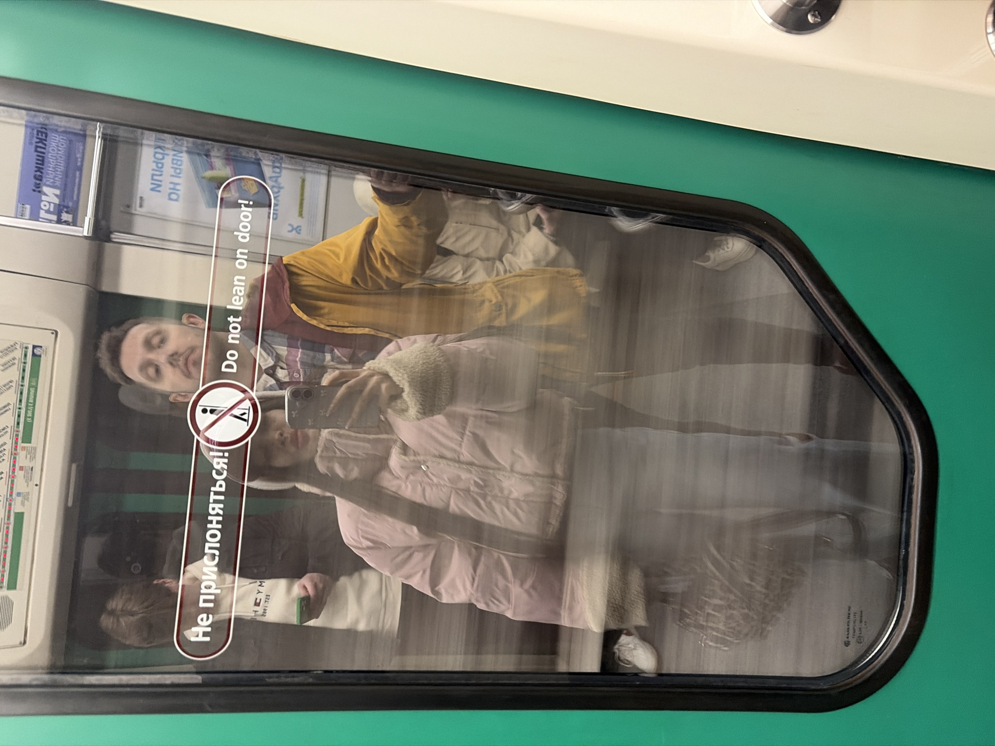 A reflective image of two people, presumably a couple, is seen through a train window. A man with short brown hair and a mustache is wearing a yellow jacket and looking to his right. A woman, partially in view, wears a light pink coat and holds a smartphone, seemingly taking a picture. The window has stickers with safety instructions in English ("Do not lean on door!") and Russian ("Не прислоняться!"). The surrounding environment appears to be the interior of a train car, with green and cream-colored panels visible. The train car has some sort of electrical access panel on the ceiling. The train car is likely to be in the city of Saint Petersburg, Russia.