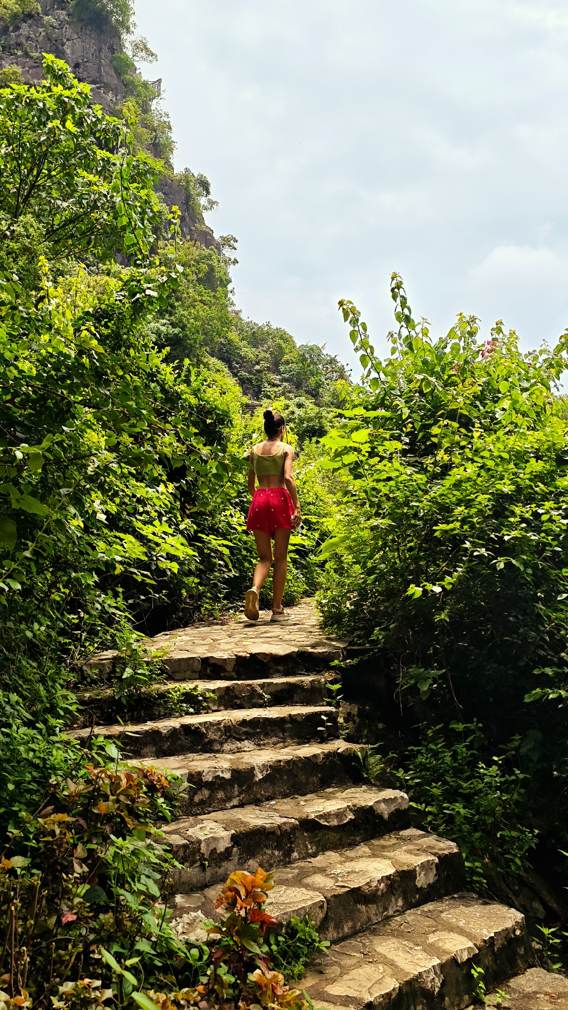 A full shot shows a woman walking up a stone staircase in Tam Thanh Ward, Vietnam. The woman is wearing a green crop top and a red skirt. She has her hair pulled back in a bun and is wearing sneakers. She is walking away from the camera, towards the top of the stairs. The staircase is made of large, uneven stones and is surrounded by lush green foliage. Trees and plants grow on either side of the stairs, and a rocky cliff face is visible in the background. The sky is cloudy.