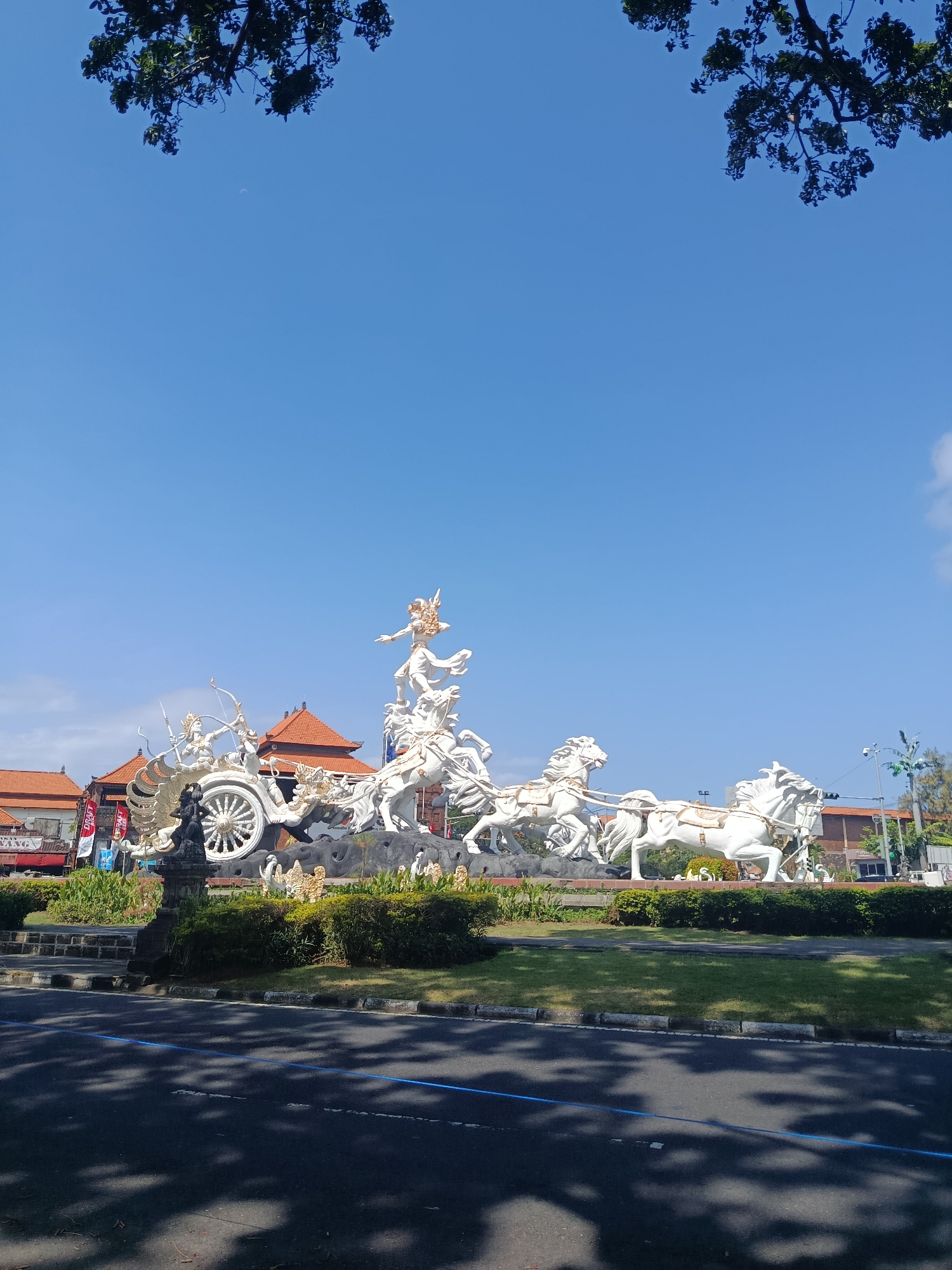 The image displays a large, white sculpture of a chariot drawn by multiple horses in Kuta, Indonesia. The sculpture is situated outdoors with a clear blue sky above. Trees with green leaves overhang from the top left of the frame. The sculpture depicts figures in dynamic poses, suggesting movement and a narrative scene. In the background, traditional Indonesian buildings with orange tiled roofs are visible, along with some greenery and street elements like traffic lights. The foreground shows a road with shadows cast by the trees, indicating it is daytime. The overall scene suggests a public monument or landmark within the city.