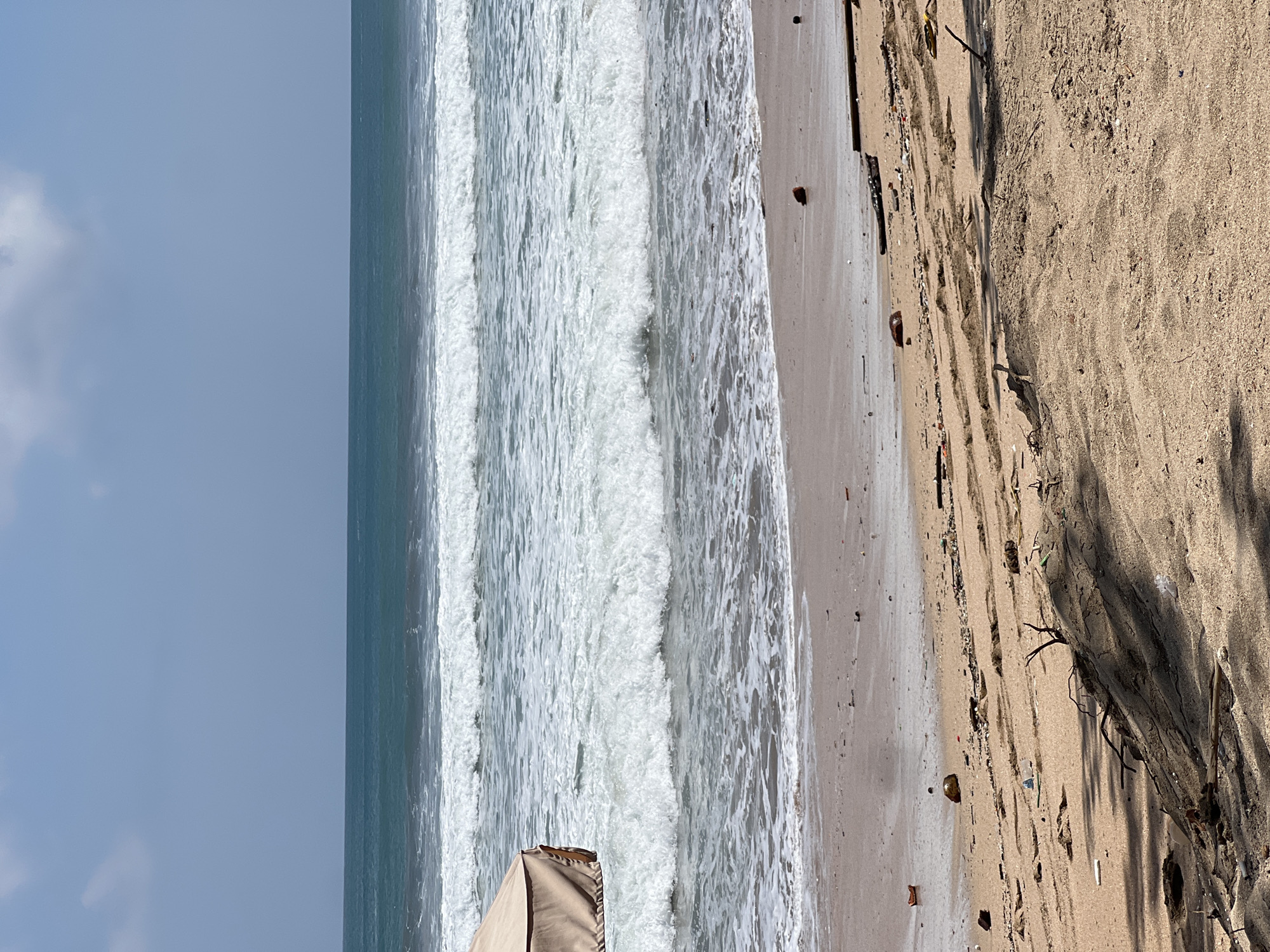 The image captures a section of Kuta Beach in Indonesia on a clear day. The scene is dominated by the ocean with several rows of white-capped waves rolling towards a sandy shore. A portion of a beige umbrella is visible on the left side, providing shade. The beach itself is littered with debris, including plastic, wood fragments, and organic matter, indicating signs of pollution. The sand is a light brown color, with some areas showing ripples from the receding tide. The sky is a pale blue with a few wispy clouds. The overall impression is of a natural beach environment impacted by human activity and the forces of the sea.