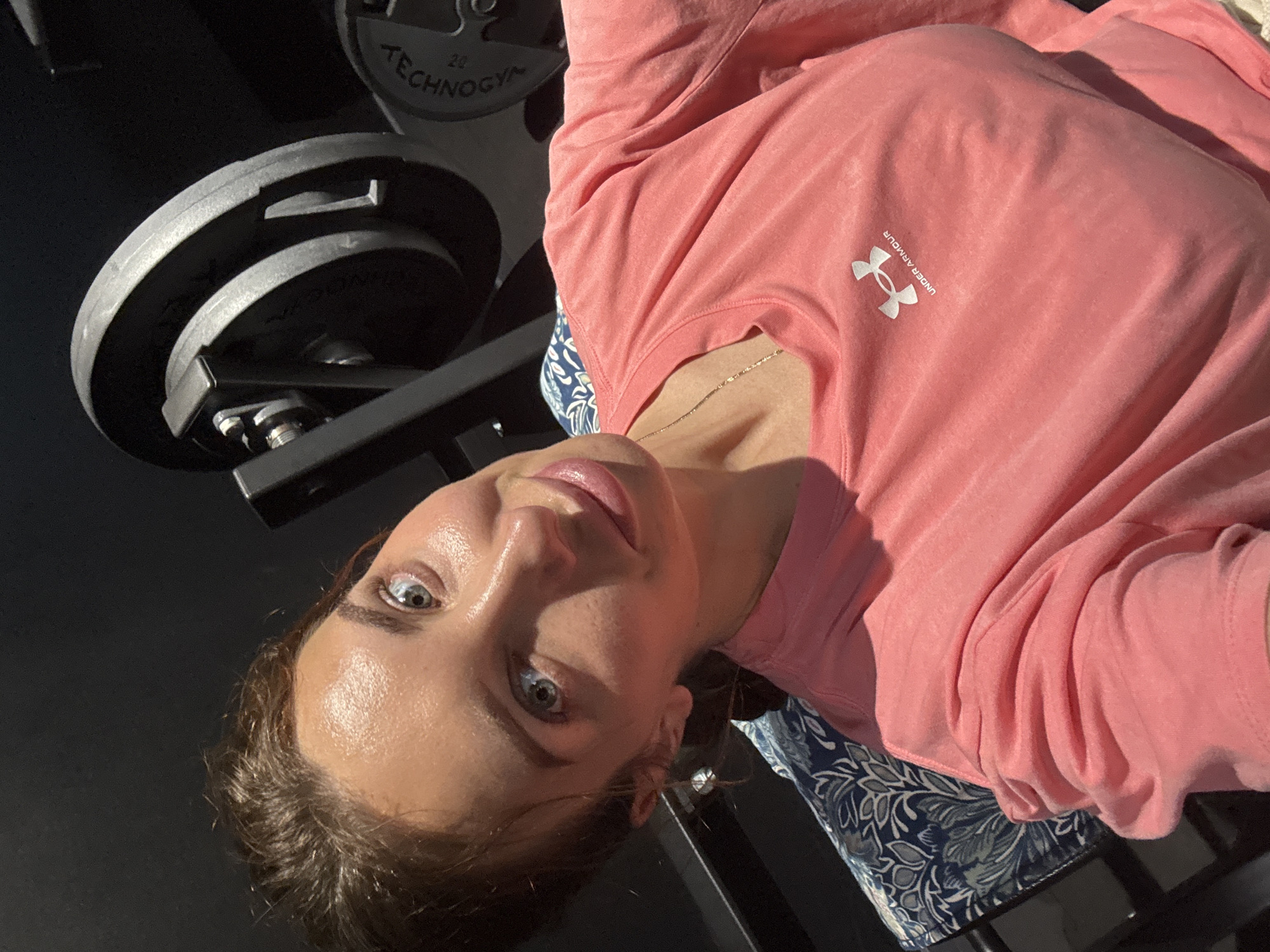The image captures a rotated, close-up perspective of a person, likely a woman, looking directly into the camera. She has light skin, blue or grey eyes, and dark hair pulled back, showing a slightly shiny or dewy face, possibly from sweat. Her lips are slightly parted, revealing a hint of a smile.
She is wearing a long-sleeved, light pink athletic shirt. On the front of the shirt, the white "Under Armour" logo, featuring the interlocking 'U' and 'A', is clearly visible, along with the brand name "UNDER ARMOUR" written vertically beside it. A delicate gold chain necklace is also visible around her neck. Below her shirt, a blue fabric with a white floral or paisley pattern can be seen, which she appears to be resting on.
The background features dark gym equipment, specifically multiple circular barbell plates stacked on what seems to be a weight machine or rack. Text visible on one of the plates includes "TECHNOGYM" and the number "20", indicating a 20 kg weight from the Technogym brand. The setting is indoors, likely a gym or a home workout space, possibly in Valencia, Spain, given the location context. Strong, natural-looking light illuminates the scene from the right, highlighting the person's face and shirt. The overall impression is that of a selfie or a photo taken during or after a workout.