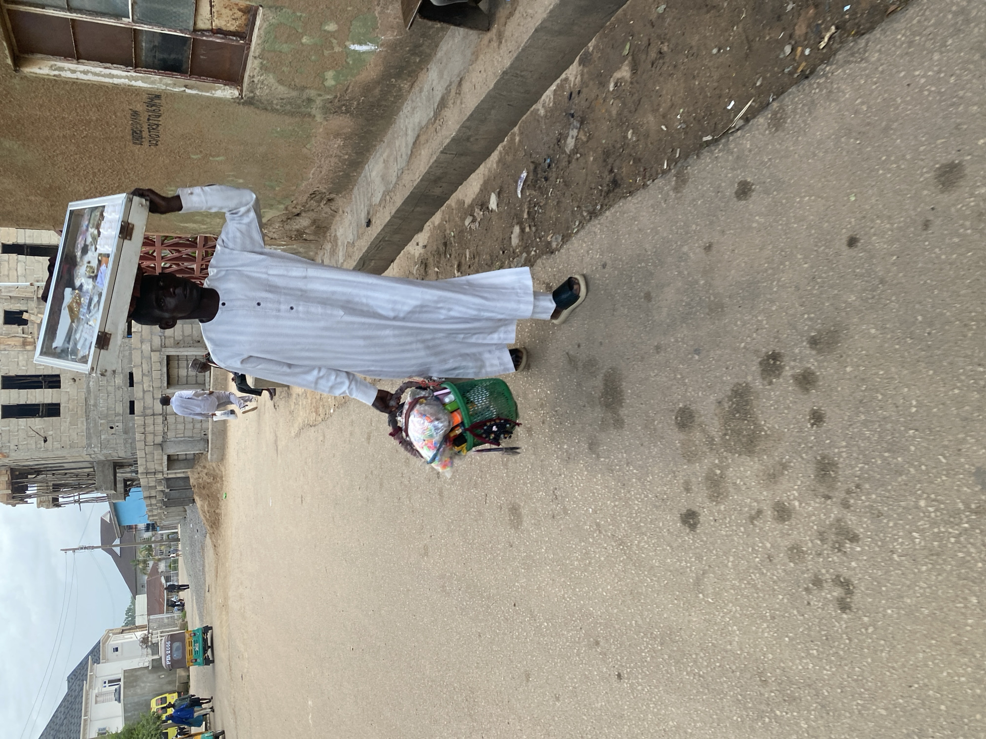 A young Black man walks along a paved street in Jos, Nigeria, carrying a white display case filled with items on his head and a green plastic basket containing various goods in his right hand. He is wearing a long, white striped garment and dark sandals. The street is lined with buildings, some of which are under construction. A yellow and green motorized tricycle is visible in the background. The sky is overcast. The scene appears to be a commercial or residential area.