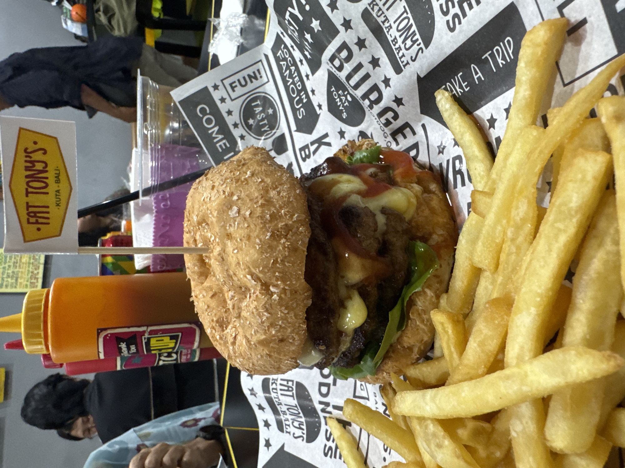 The image shows a burger and fries meal at Fat Tony's in Kuta, Bali. The burger features a textured bun and is topped with cheese, greens, and a sauce that appears to be ketchup. A Fat Tony's flag is stuck into the burger. Alongside the burger is a generous serving of golden fries. A ketchup and mustard bottle are present near the burger. The meal rests on branded paper with "Fat Tony's" printed on it, also mentioning "Kuta-Bali" and phrases like "Take A Trip" and "Located in Famous." A person wearing a dark top can be seen in the background, seemingly at a table in the restaurant.